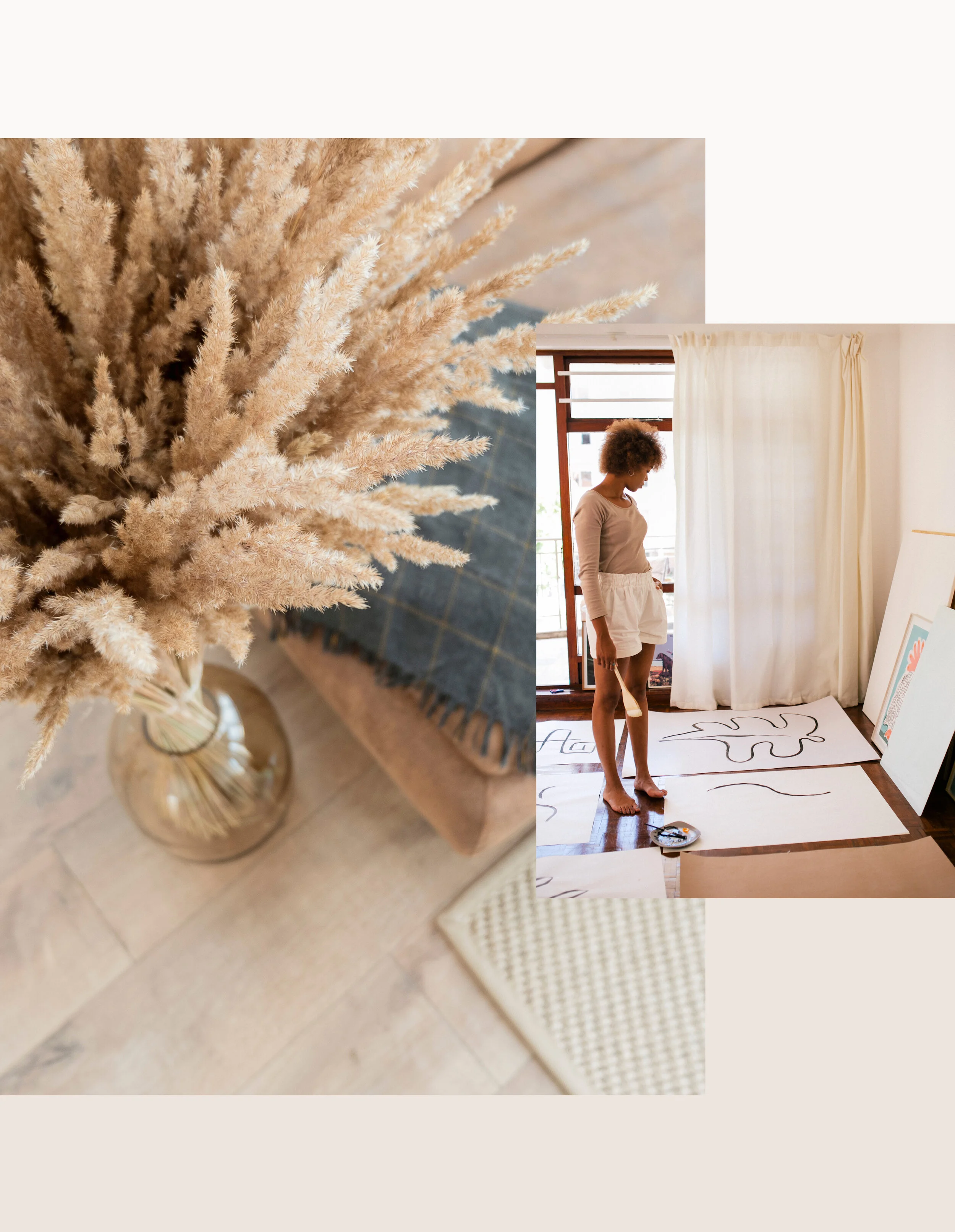 A close-up of beige dried pampas grass in a glass vase on a wooden floor, with a beige textured mat nearby. Behind, a woman is painting large abstract artworks on paper spread on the floor, near a window with white curtains.