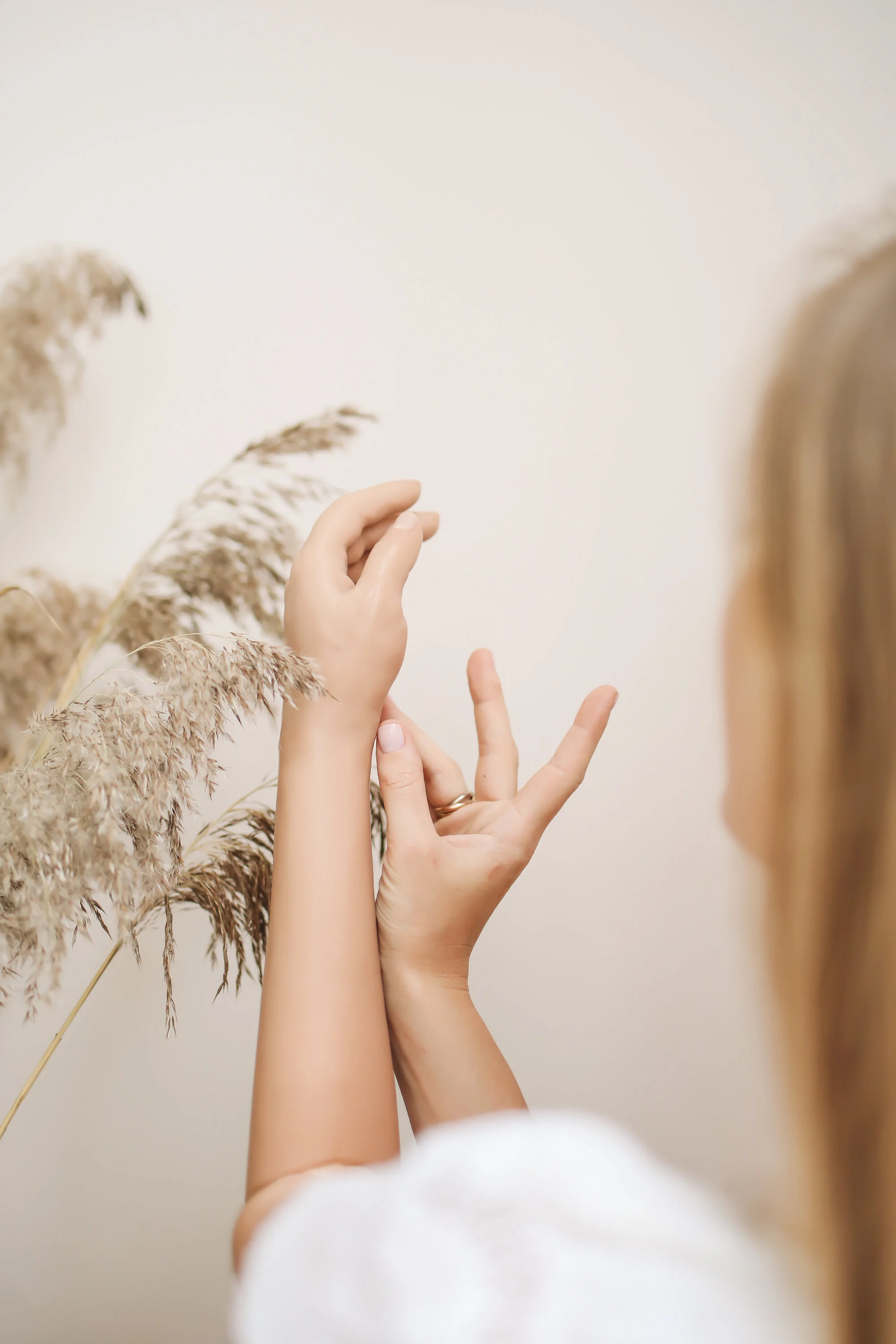 Person performing a hand gesture near tall beige decorative grasses against a plain white wall.