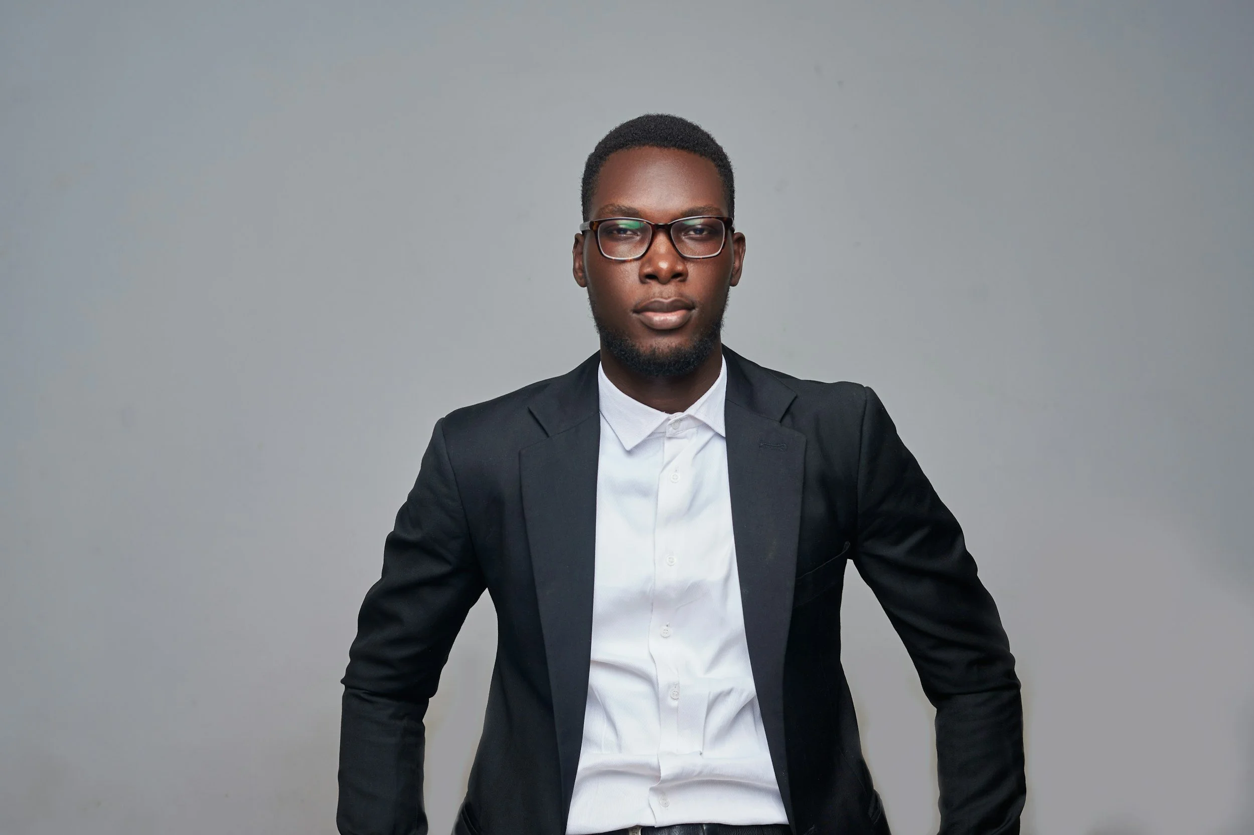 A young man dressed in a black suit and white shirt standing against a plain gray background.