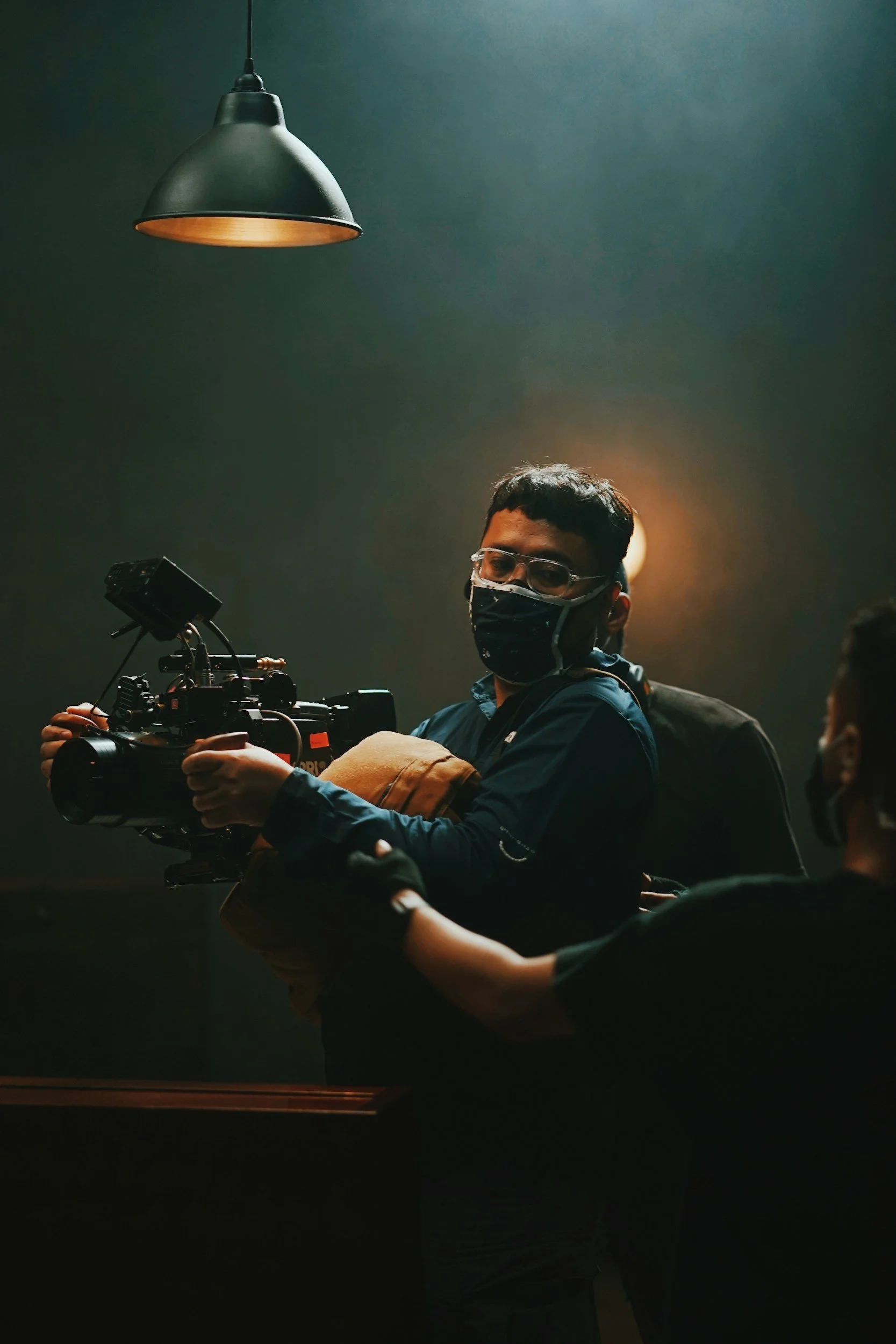 Film crew member wearing a face mask and glasses handling a professional camera, with a spotlight overhead and another person assisting.