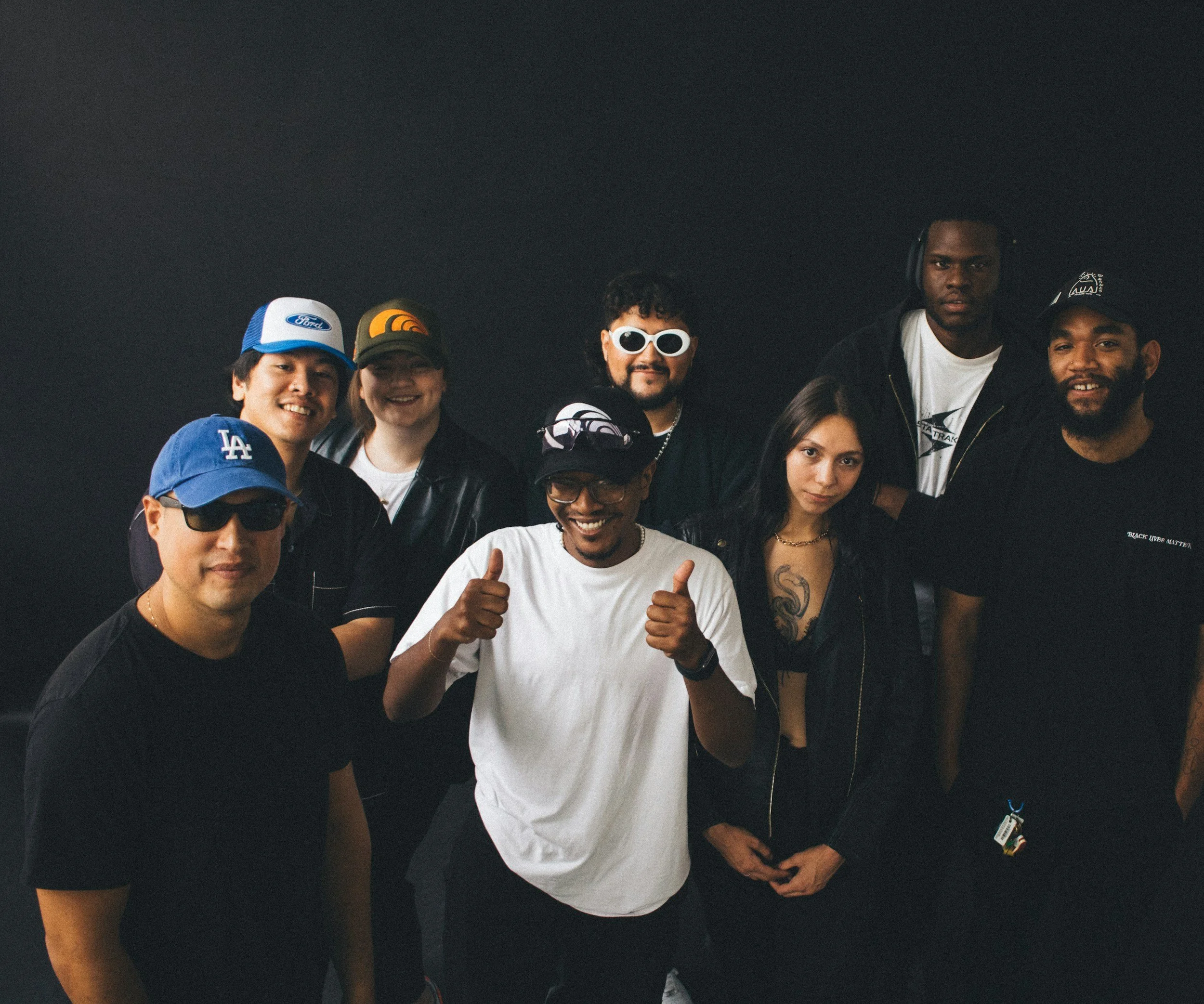 A diverse group of nine people standing together against a dark background, smiling and posing for the photo. Some are wearing caps and sunglasses, and one person is giving thumbs up.