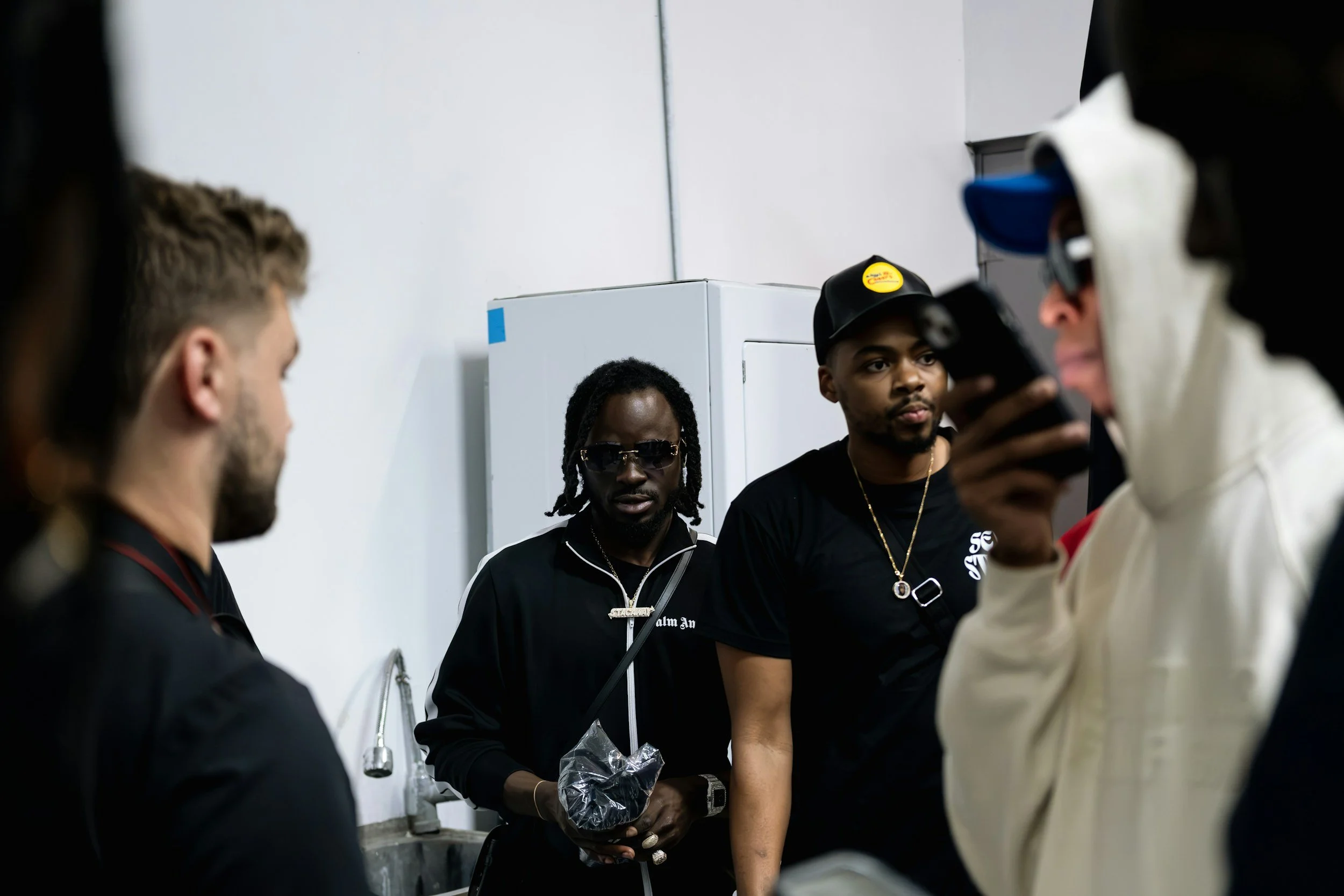 Group of young men gathered indoors, some wearing dark clothing, one in sunglasses, with a white wall and a white appliance in the background.