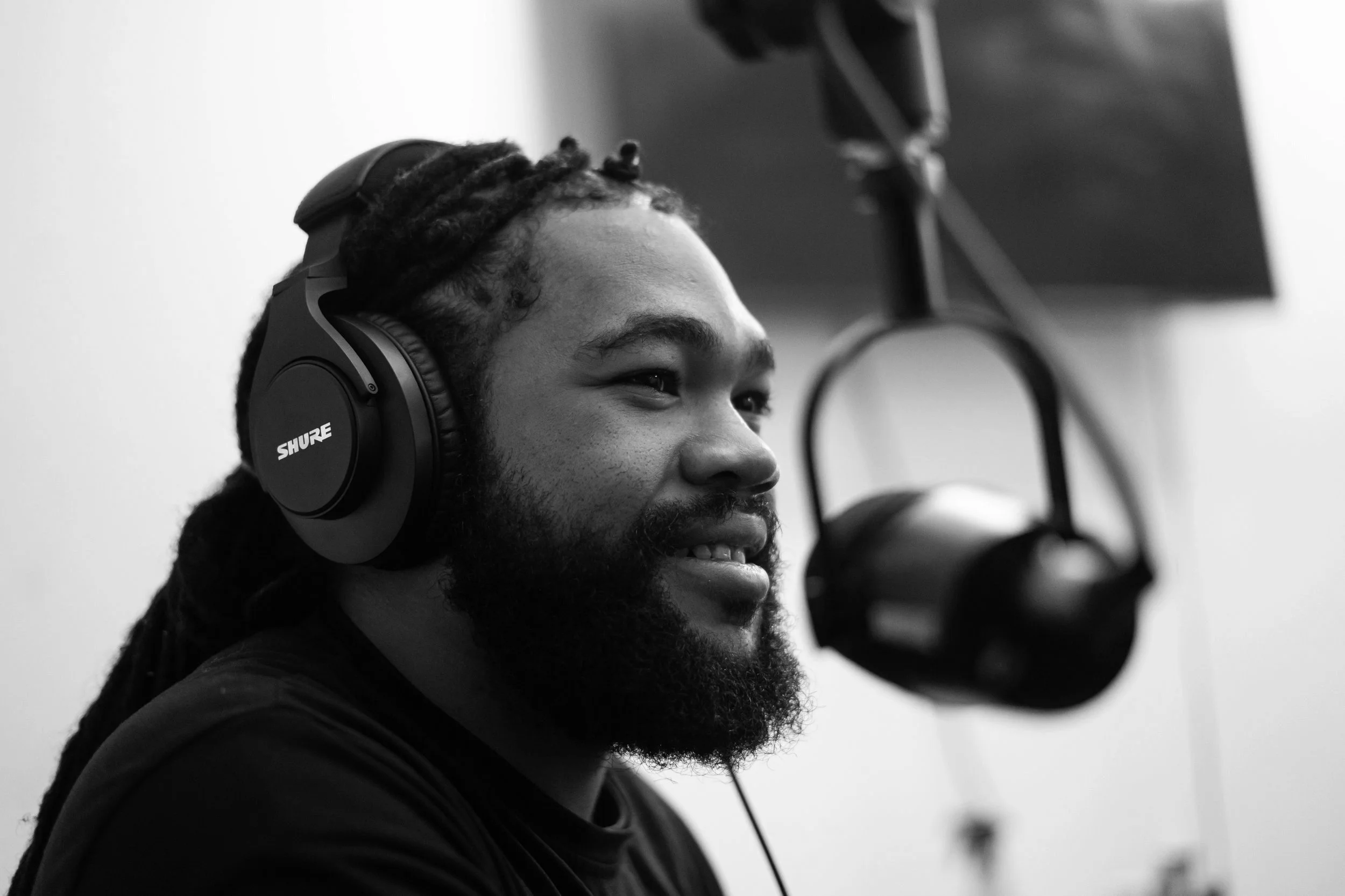 Black and white image of a man with dreadlocks and a beard wearing headphones, sitting in front of a microphone.