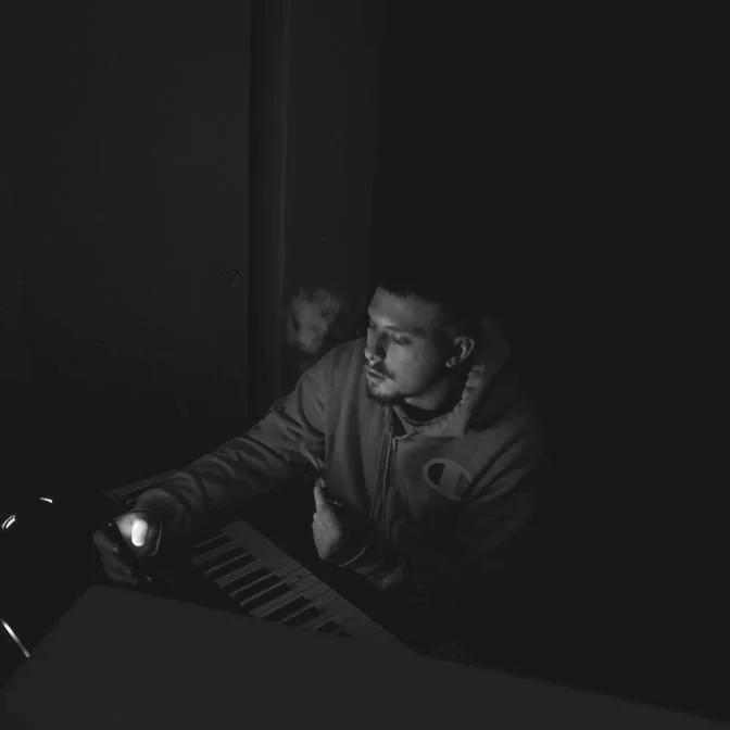 A young man sitting in a dark room at a desk, holding a mobile phone with one hand, with a keyboard in front of him, and smoking a cigarette.