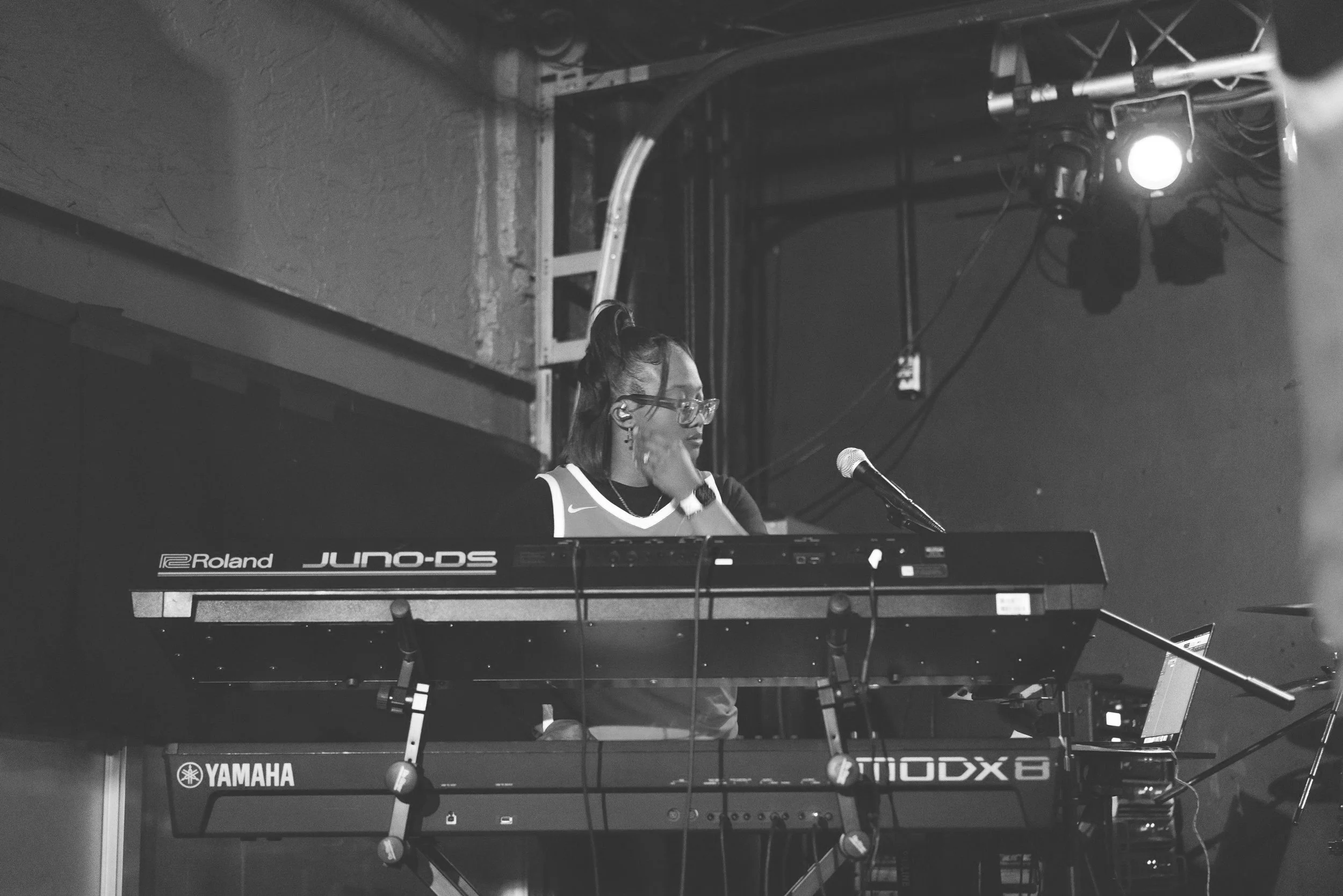 A person with glasses and earrings standing behind two electronic keyboards, Roland and Yamaha, on a stage with stage lights, cables, and a laptop, as they work on music, in a black and white photo.