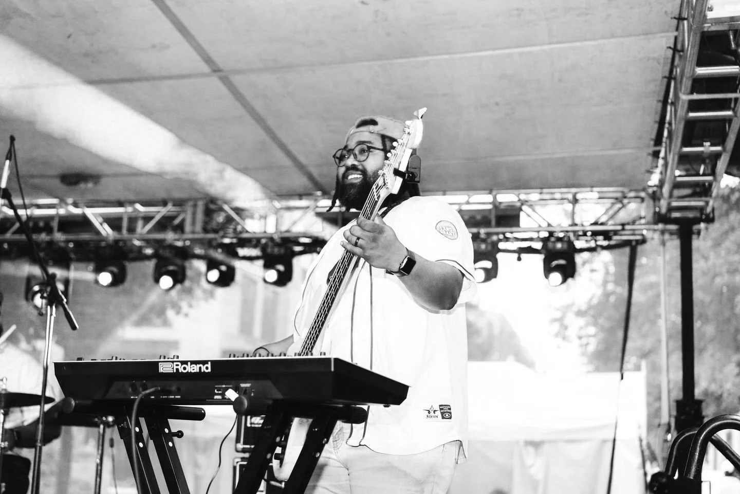 A musician with glasses, a beard, and a backward cap playing a bass guitar and smiling while standing behind a Roland keyboard at an outdoor concert stage.