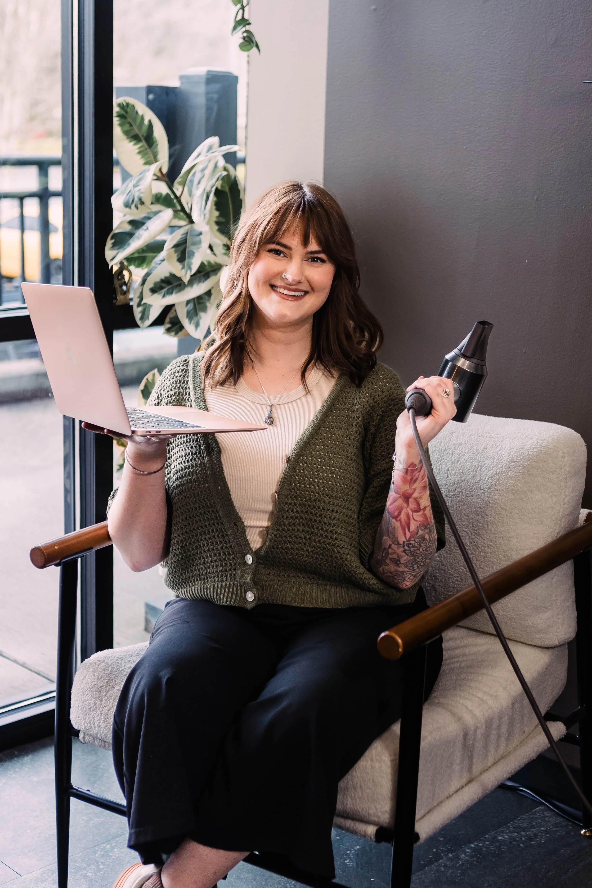 A cozy workspace with a white shelving unit filled with jars of candy and supplies, a pink plush chair, a fluffy stool, and a desk with a lamp. In the background, there are pink and grey armchairs with cushions and a gold table.