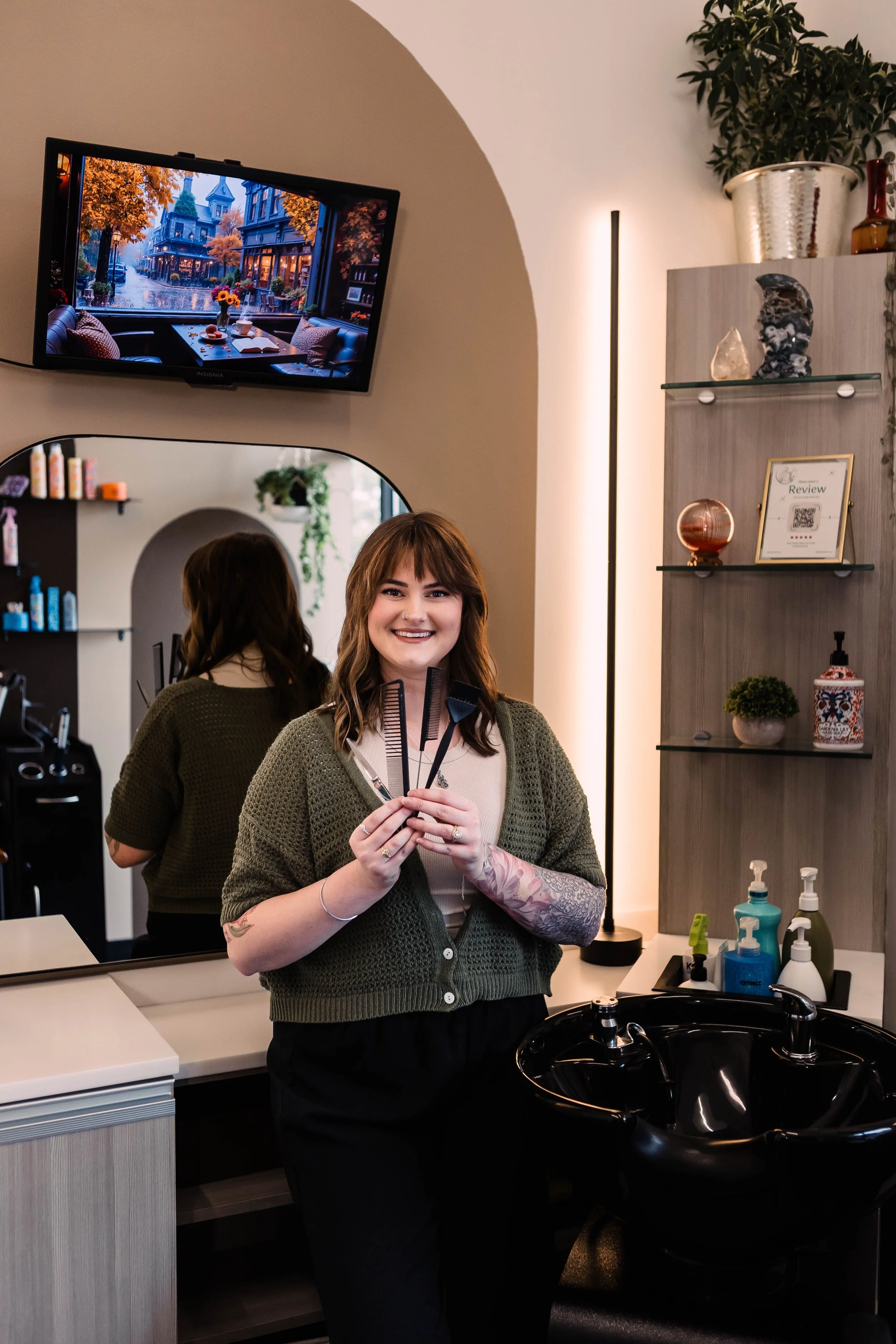 A smiling woman holding hairstyling tools in a salon, standing in front of a mirror with shelves holding hair products, a hair washing sink, and a wall-mounted TV showing an outdoor scene.