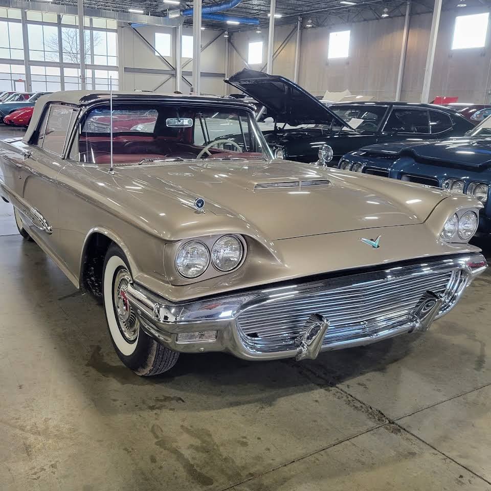 A vintage gold-colored car with dual headlights, a chrome front grille, and white-wall tires, parked inside a spacious showroom.