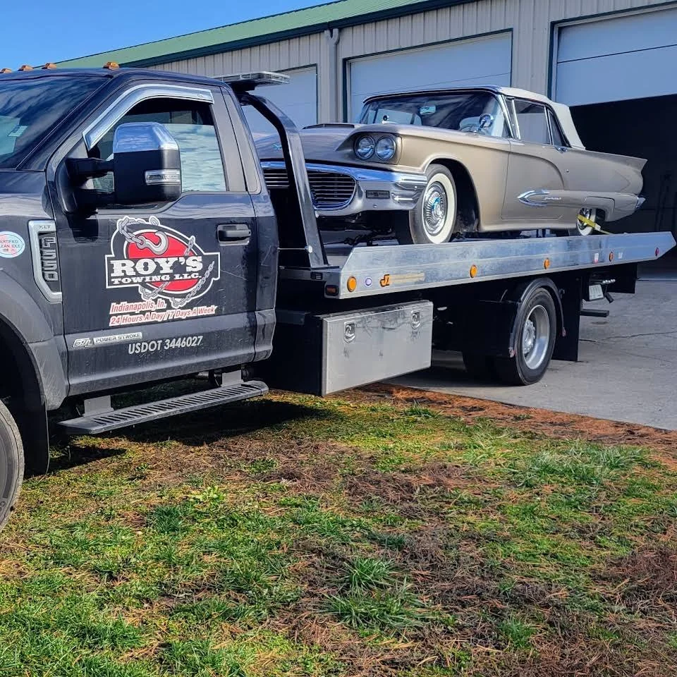A flatbed tow truck with a vintage beige and white car loaded on it, parked outside a building.