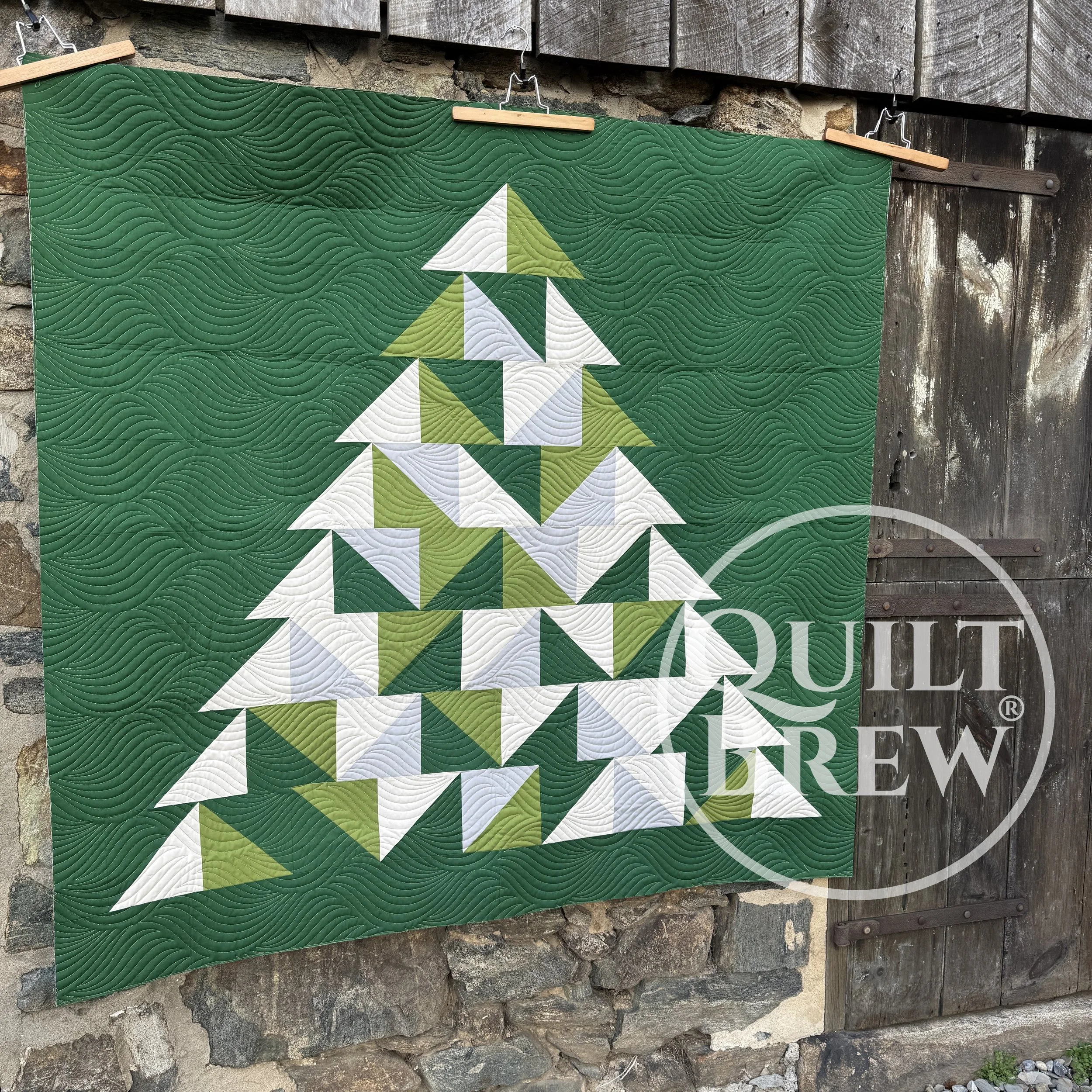 Woman with glasses holding up a quilt with a Christmas tree pattern made of white and green triangles on a dark green background outdoors near a stone and wooden barn.