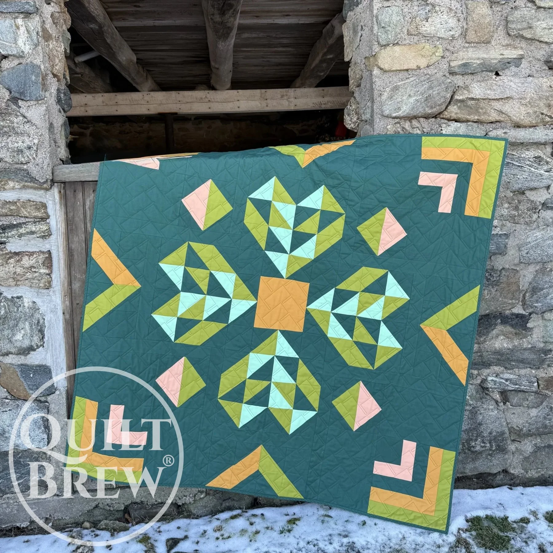 A green quilt with a center block shaped like a heart is draped over a stone barn window.