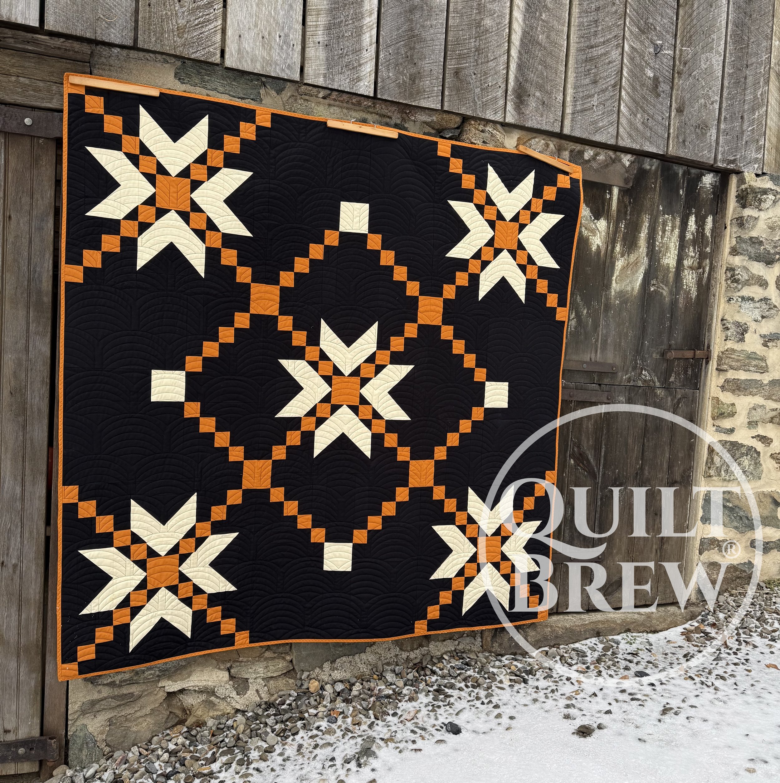 A quilt with a black background and white and orange star patterns hanging outside on a wooden and stone wall, with snow-covered ground below.