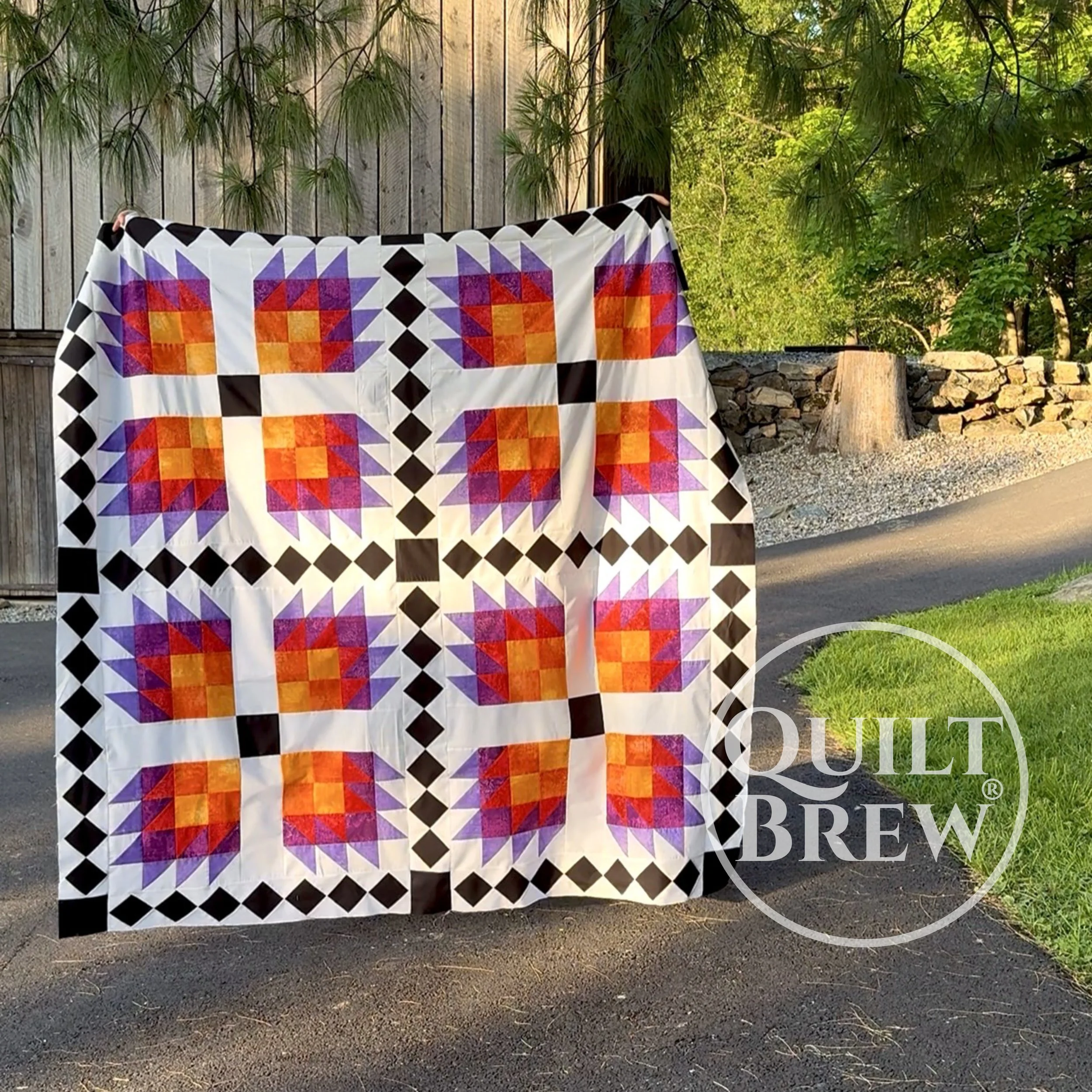 A colorful quilt with bear paw blocks featuring orange, pink, purple, and red hues, hangs outdoors. The background includes a stone barn, green trees, and a paved path with a grassy area.