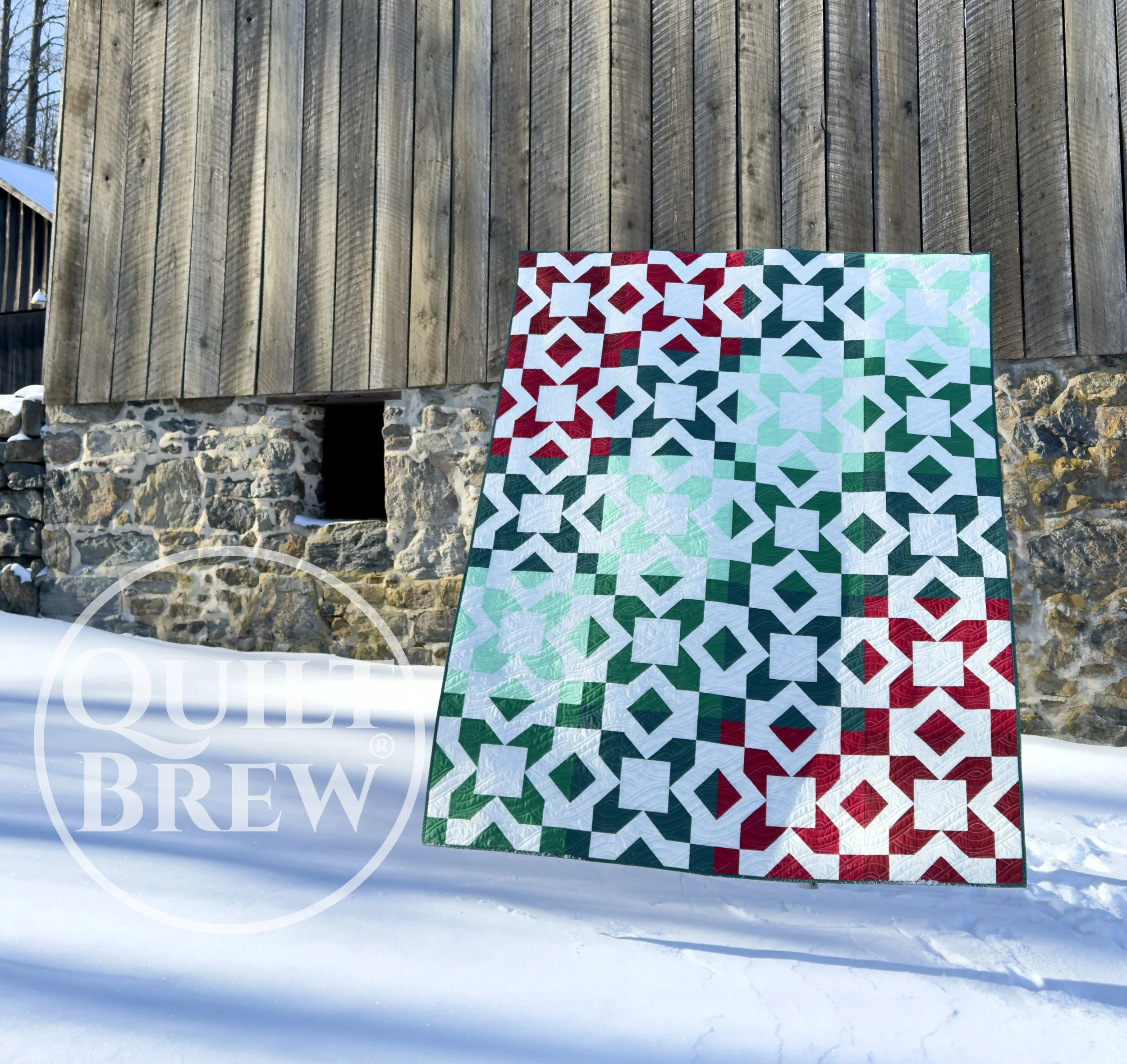 A quilt with a geometric pattern in red, white, green, and light mint colors hanging outdoors in a snowy setting, against a wooden and stone wall background.
