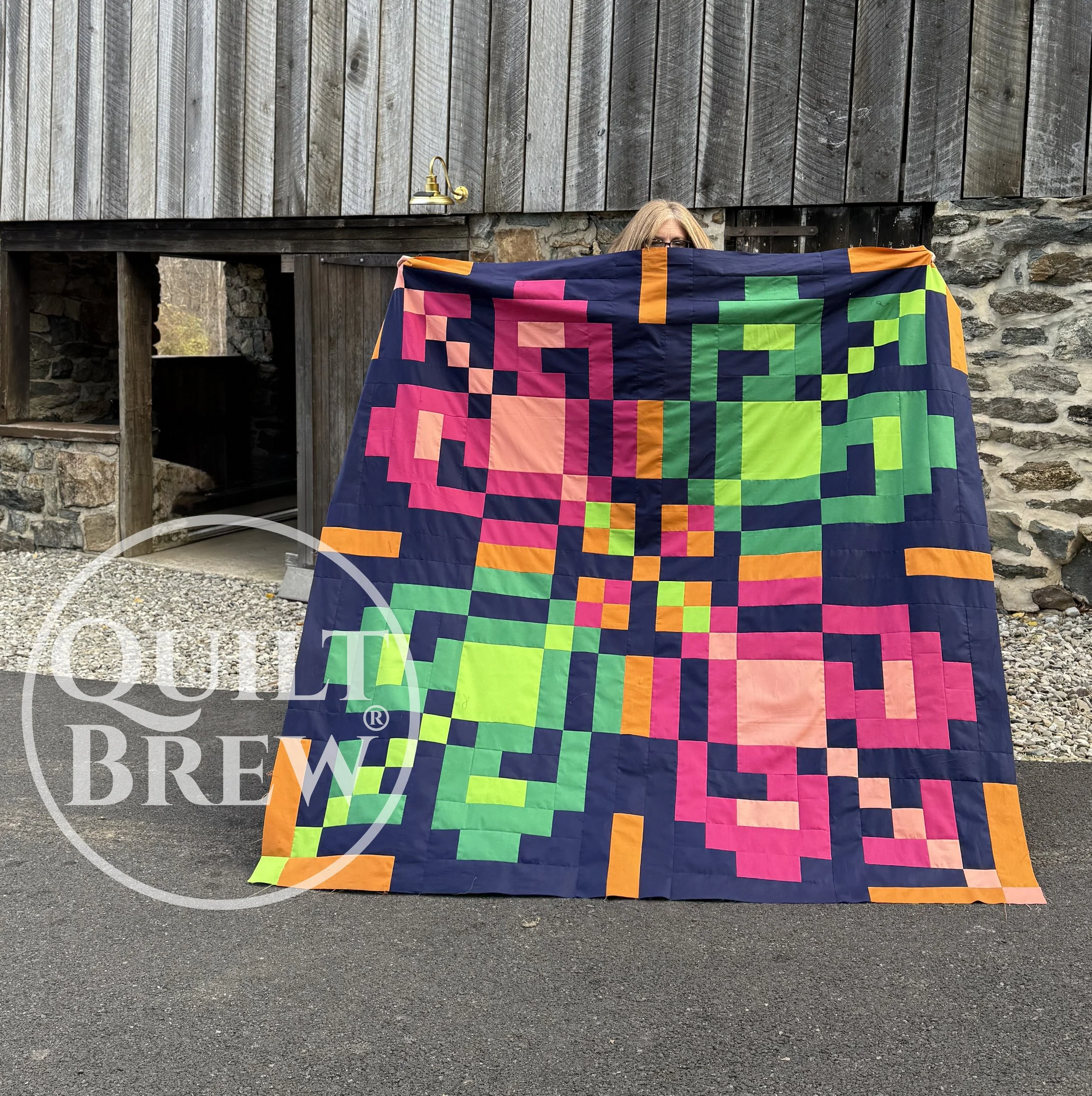 Person holding colorful quilt with pink, green, orange, and blue geometric pattern. The background features a rustic stone and wooden barn.