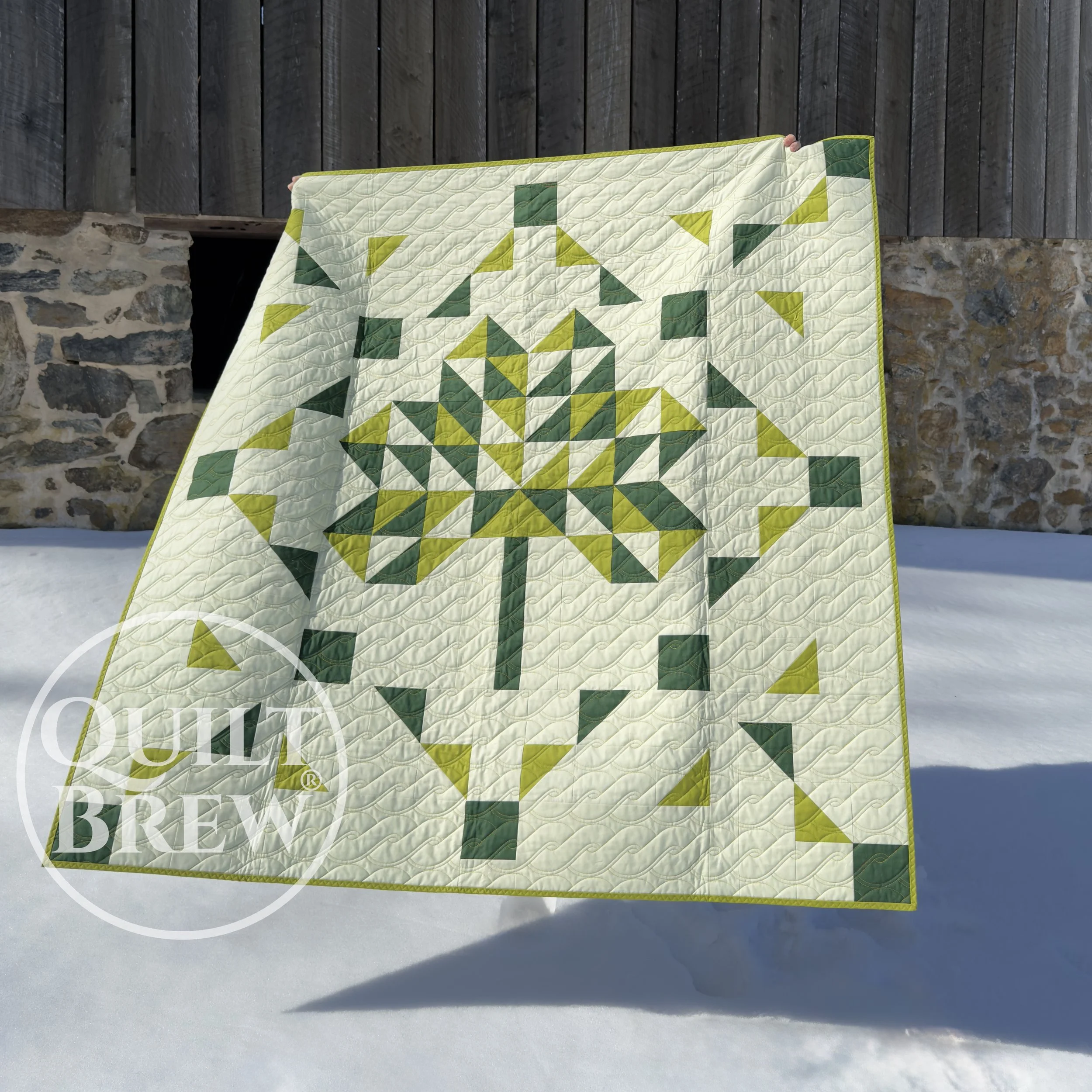 A quilt with a geometric pattern resembling a shamrock in shades of cream, emerald and sea green, hangs outside near a stone and wooden barn.