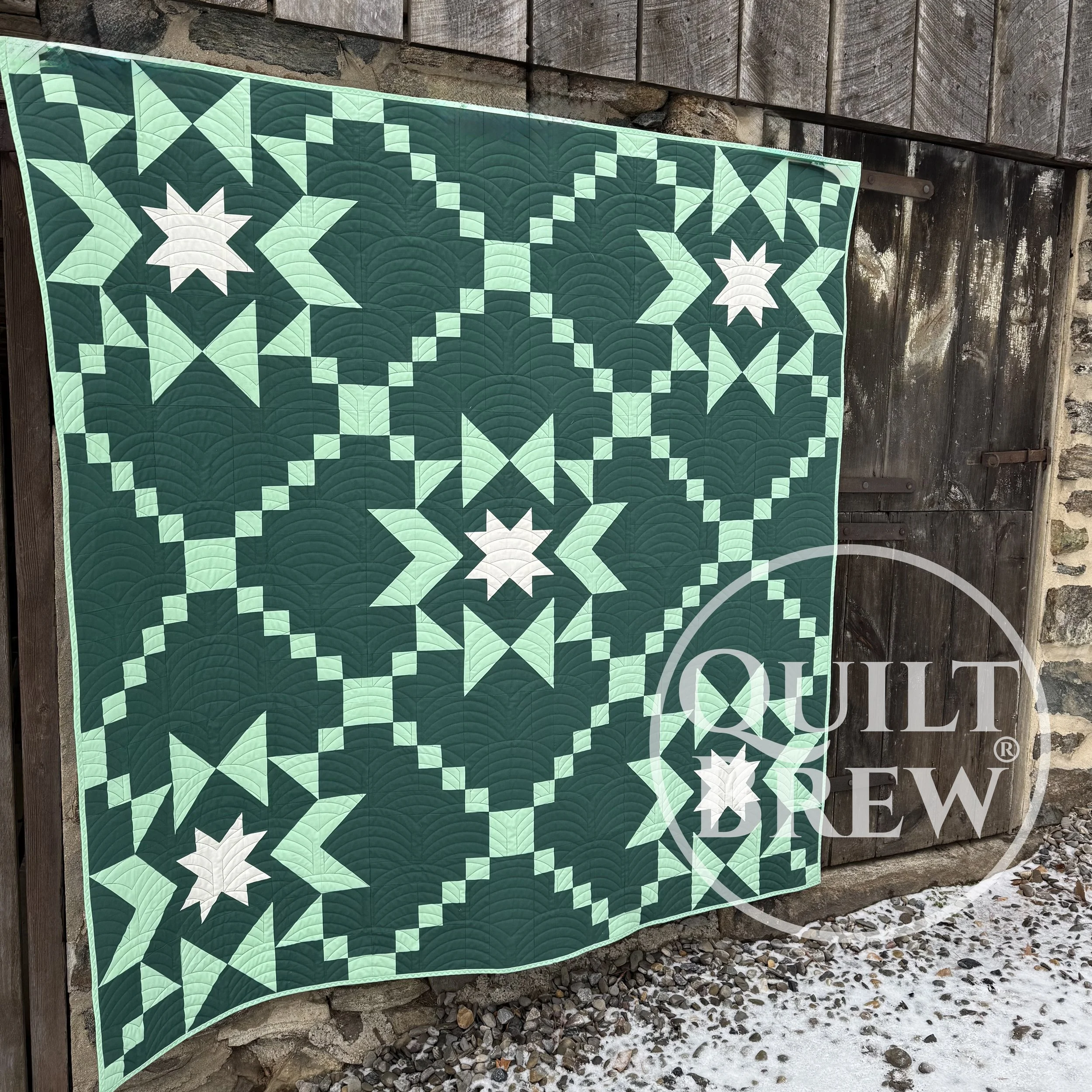 A quilt with a white, sea green and dark green star pattern hanging on a wooden wall outside, partially covering a stone wall and a door, with snow on the ground.