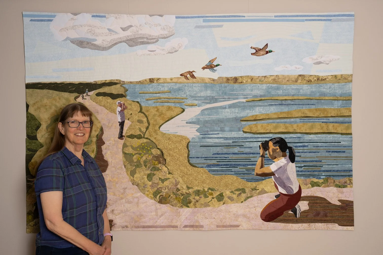 Artist Joan Sowada standing in front of art quilt titled Bolsa Chica Trails.