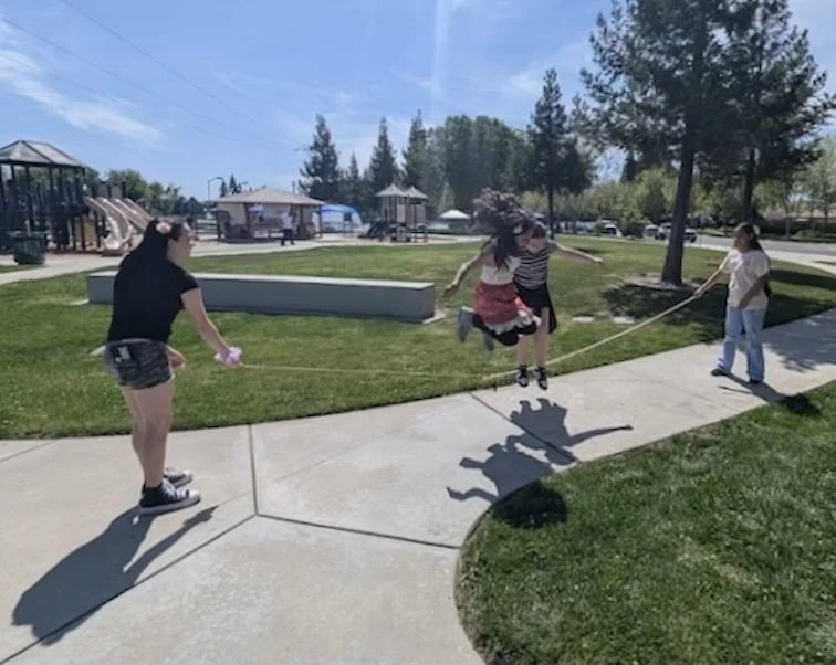 Two girls jump rope. Two older girls are on the ends moving the rope. They are at a park. Blue sky and good light and shadows.