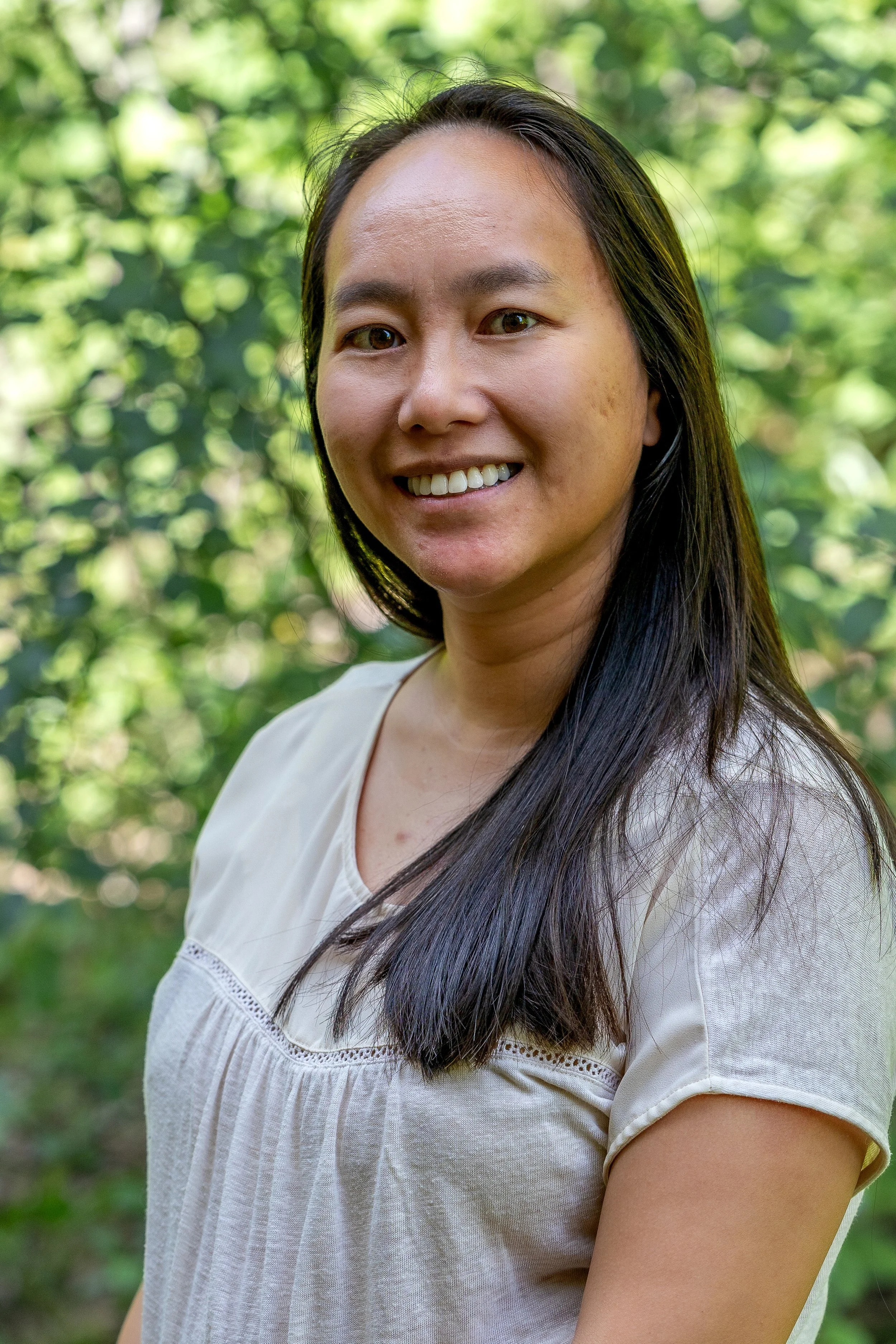 A woman with long black hair smiling outdoors in a green wooded area.
