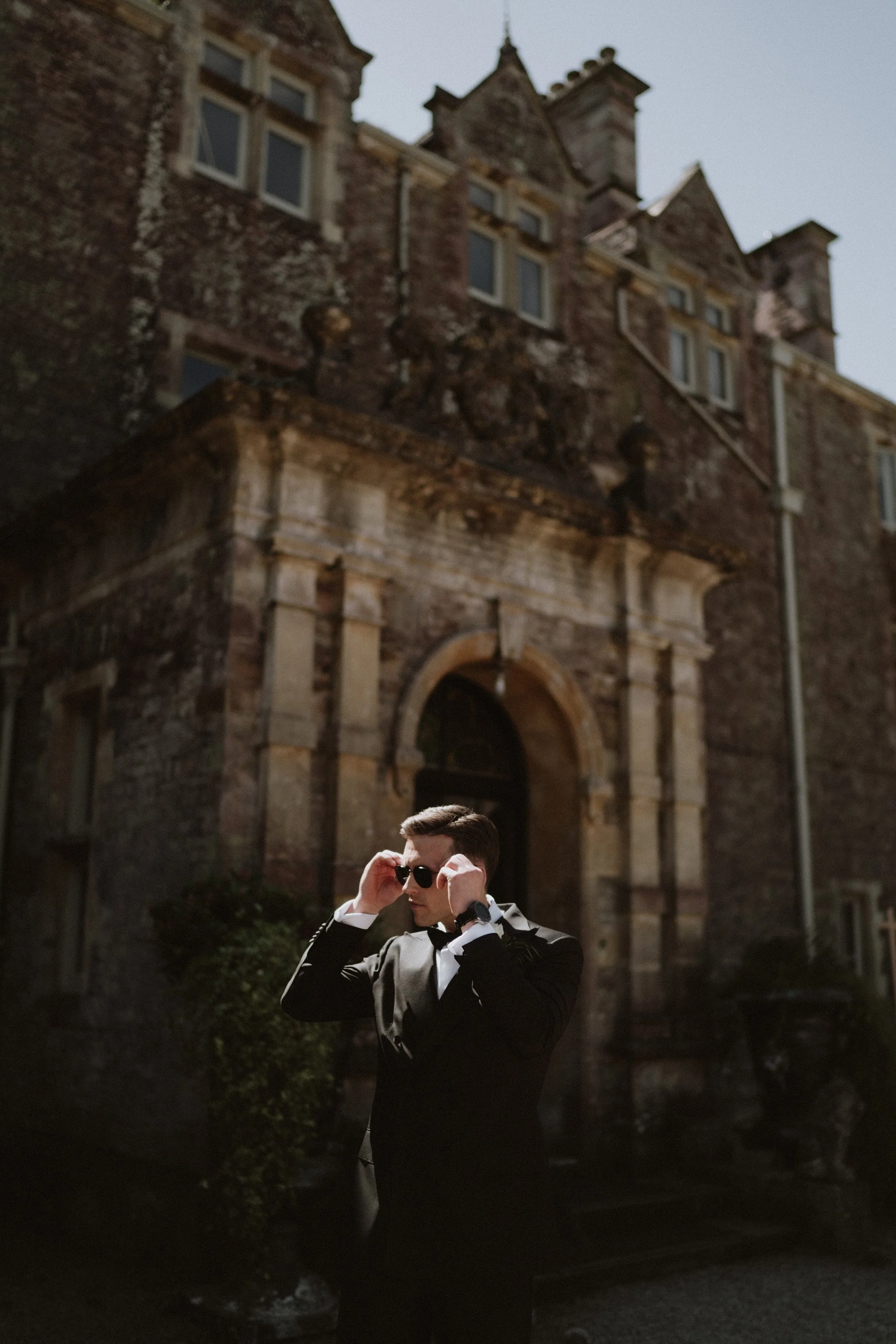 A man in a tuxedo and sunglasses adjusting his glasses standing in front of a historic stone building during the daytime.