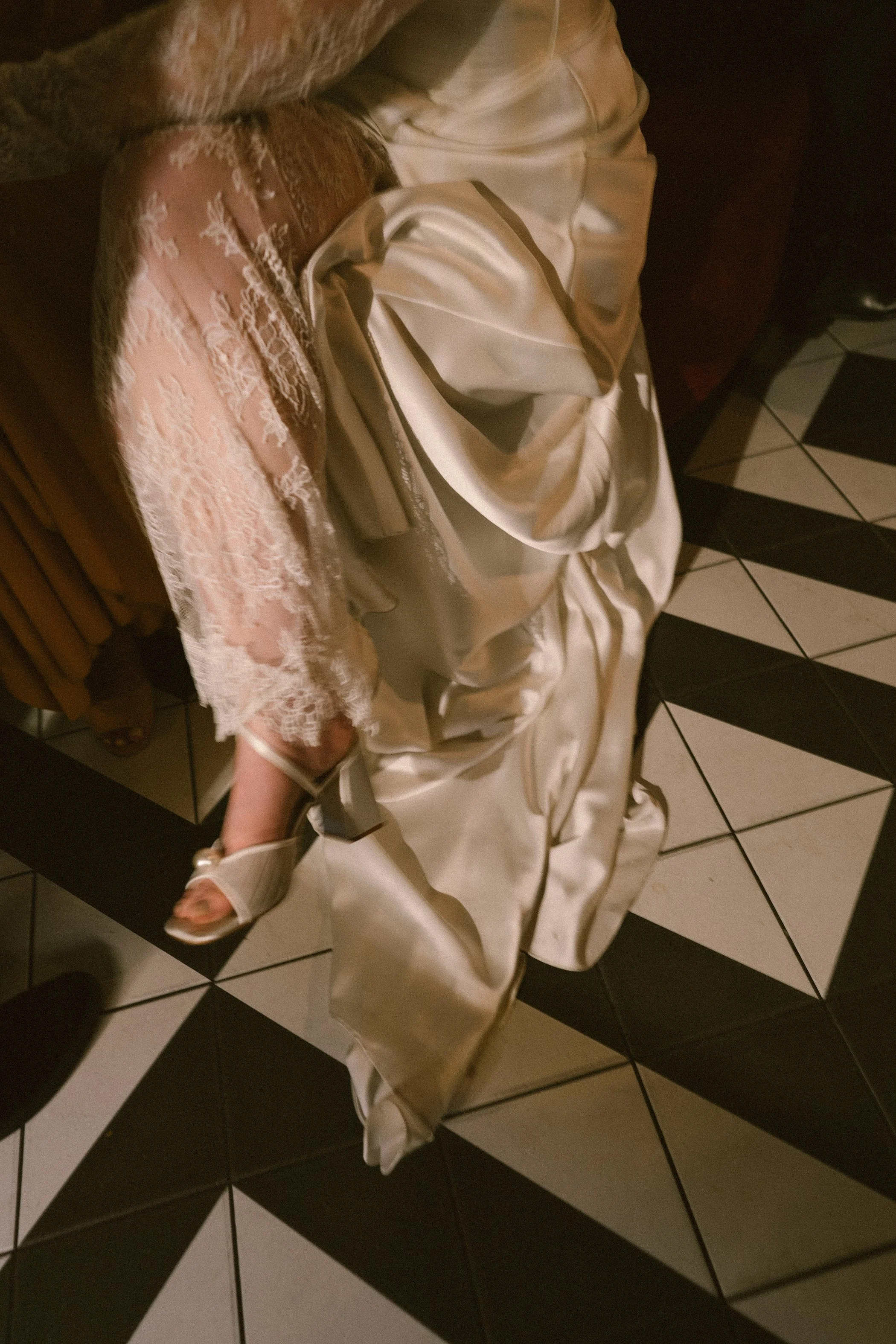 A person wearing a cream-colored satin wedding dress and silver high heel shoes sitting on a black and white tiled floor.