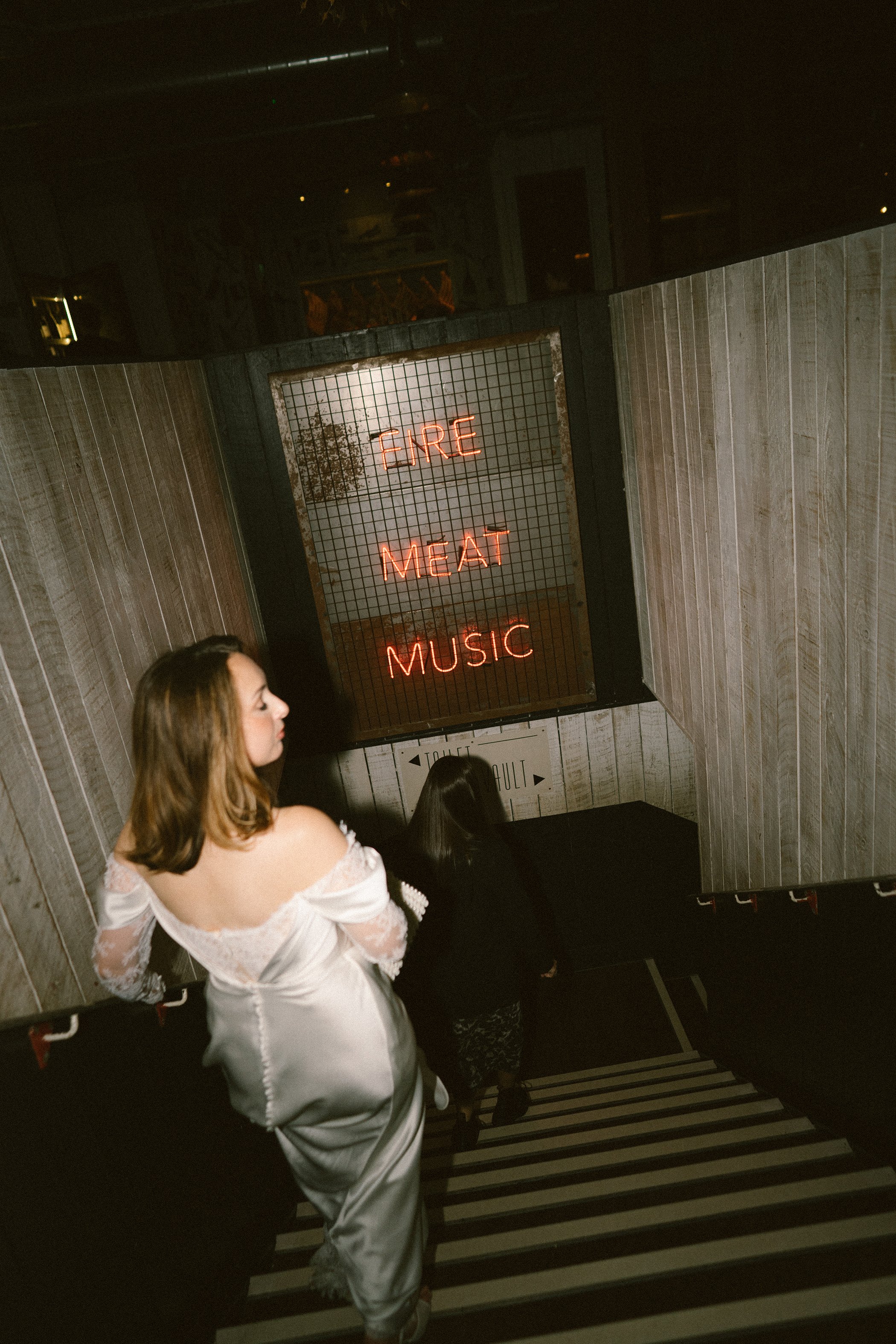 A woman with wavy brown hair in a white off-shoulder dress standing near the top of a staircase, with a neon sign reading 'FIRE MEAT MUSIC' mounted on a wire grid on the wall above her.