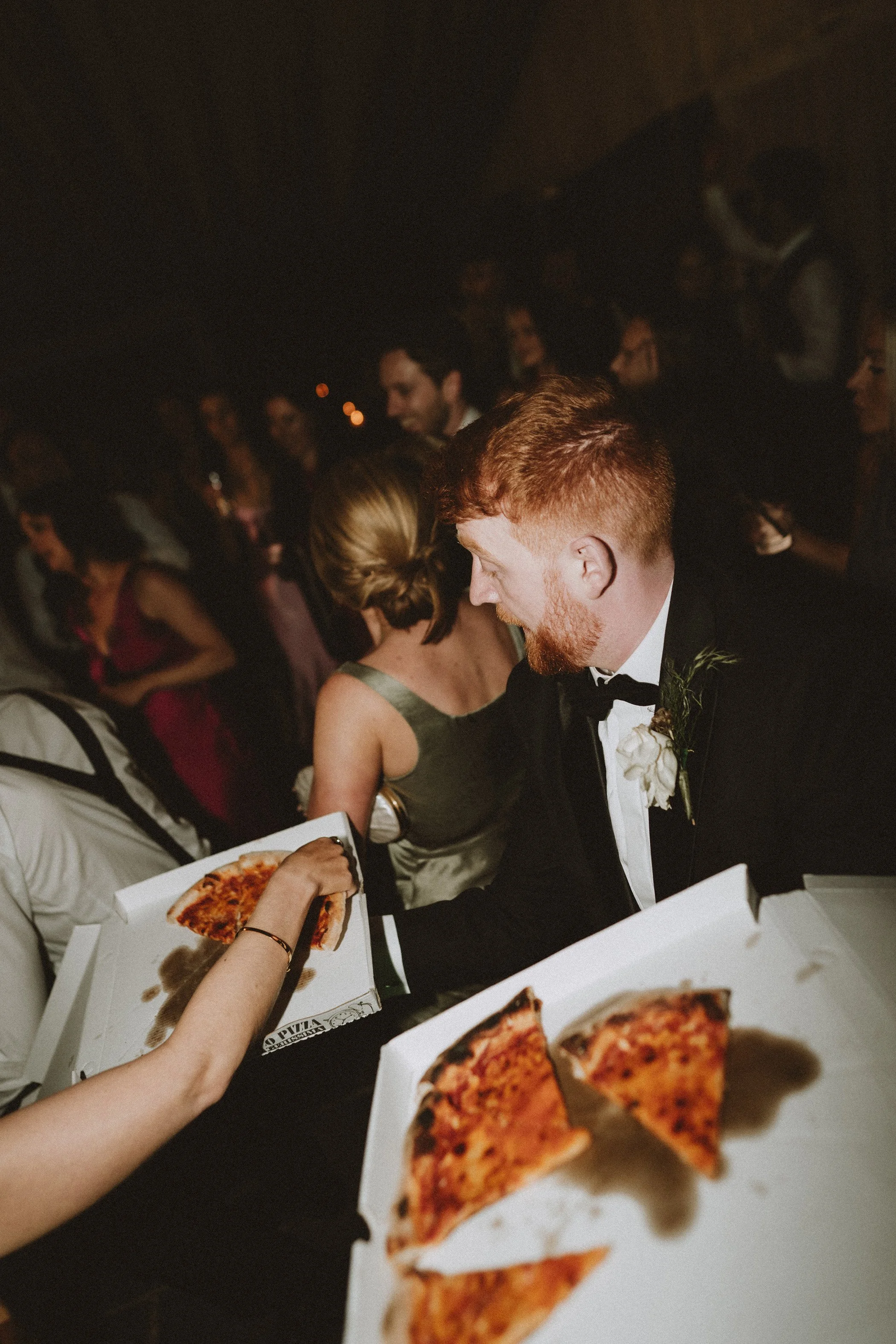A man in a tuxedo with a boutonniere receives a slice of pizza from a hand reaching out in a crowded party setting.