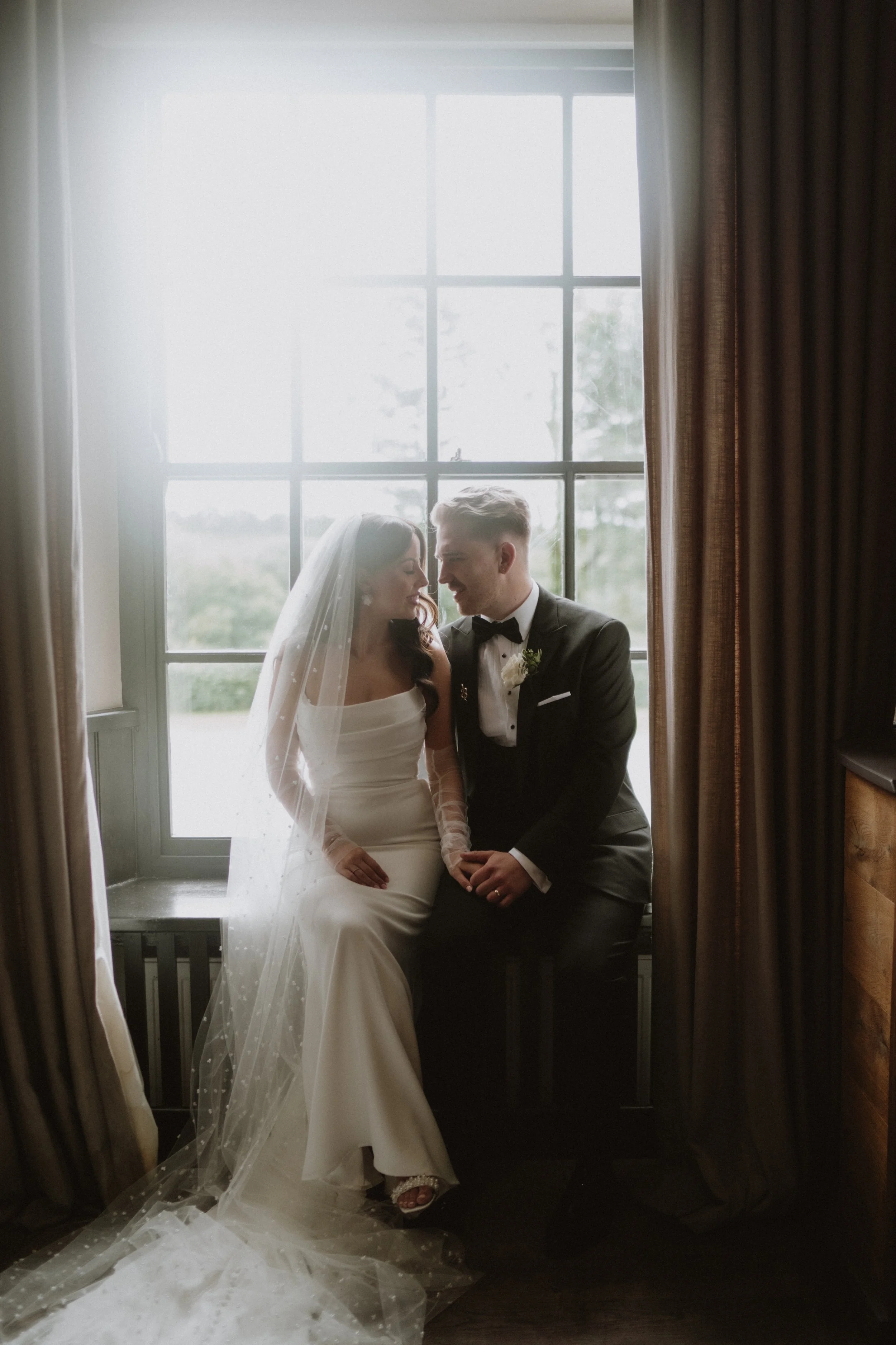 Bride and groom sitting together on a window sill, holding hands, with the bride wearing a white wedding gown and veil, and the groom in a black tuxedo, inside a room with large window and curtains. south wales uk wedding