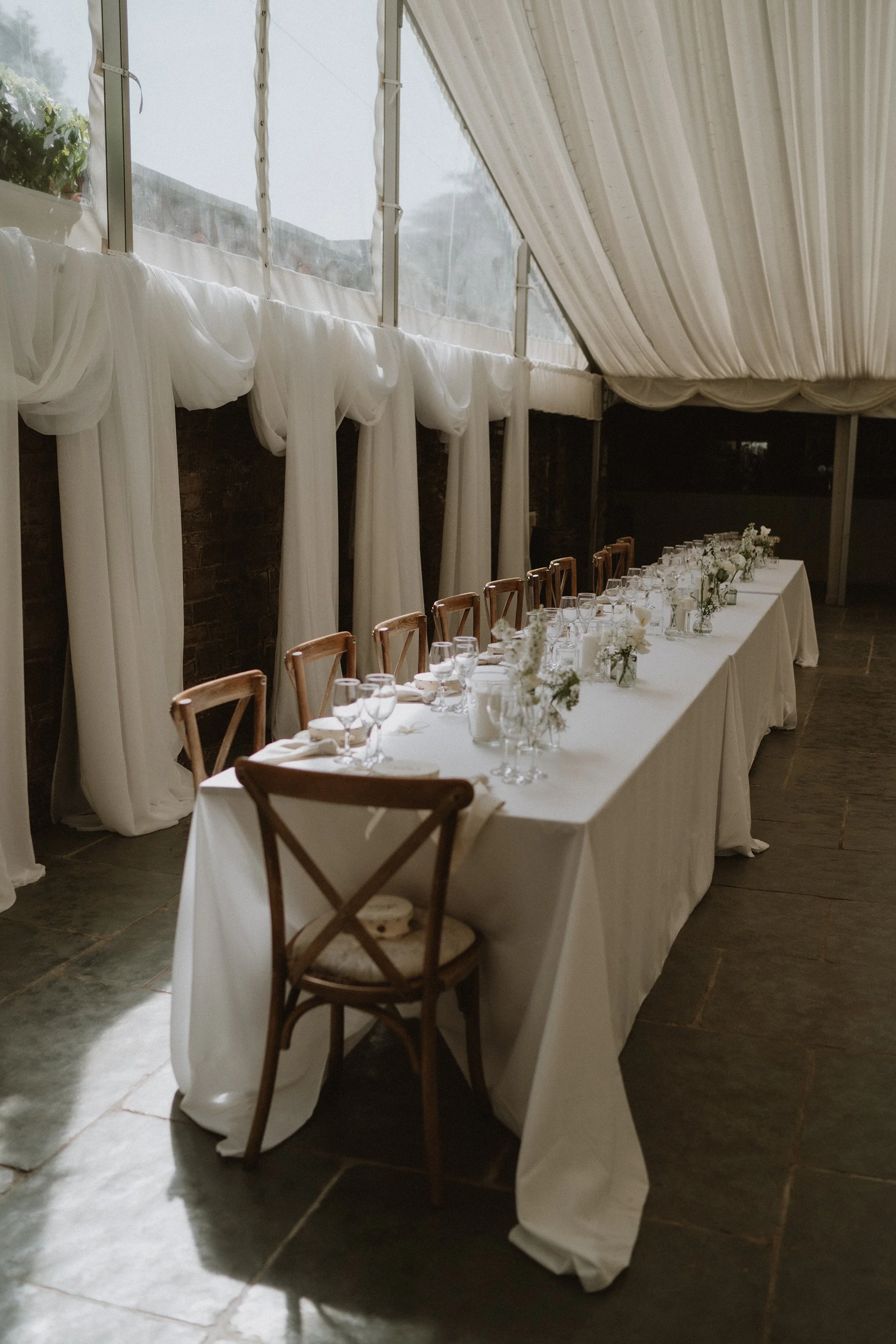 Wedding reception table with white tablecloths, glassware, and floral centerpieces inside a tent with white drapes.