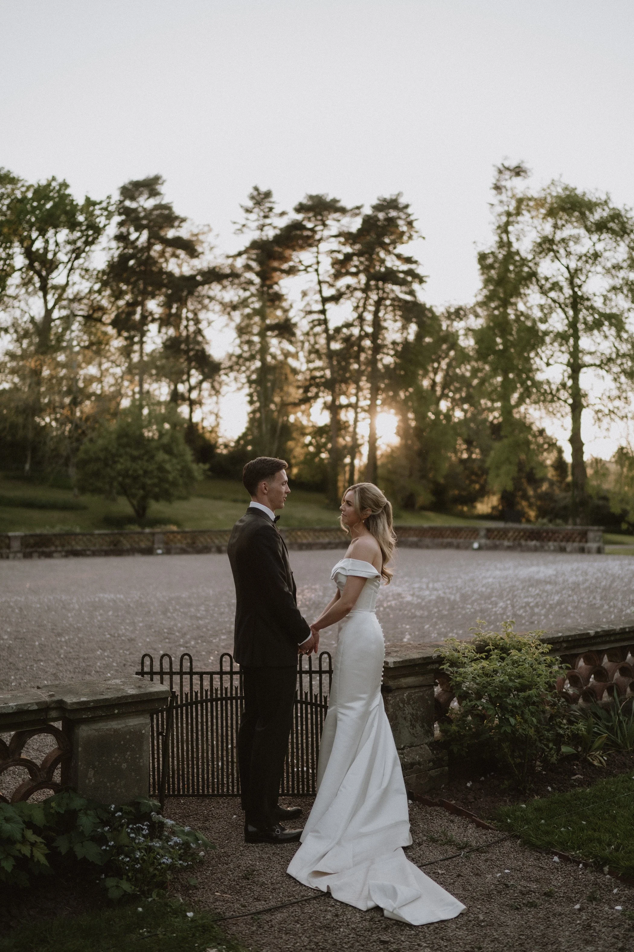 A bride and groom holding hands and looking at each other outdoors during sunset, with trees and a park in the background.