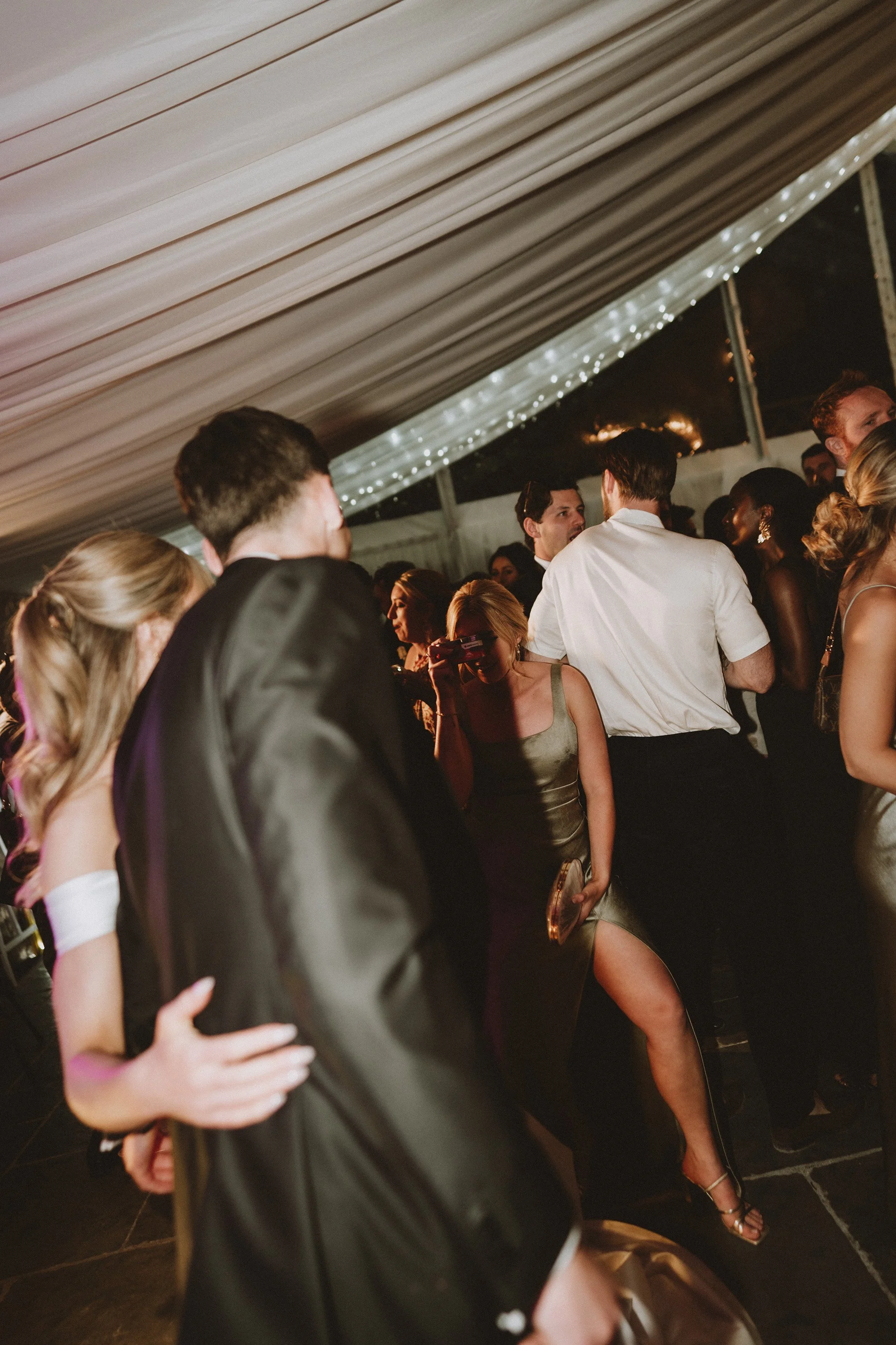 People dancing and socializing at a party under a decorated tent with string lights.