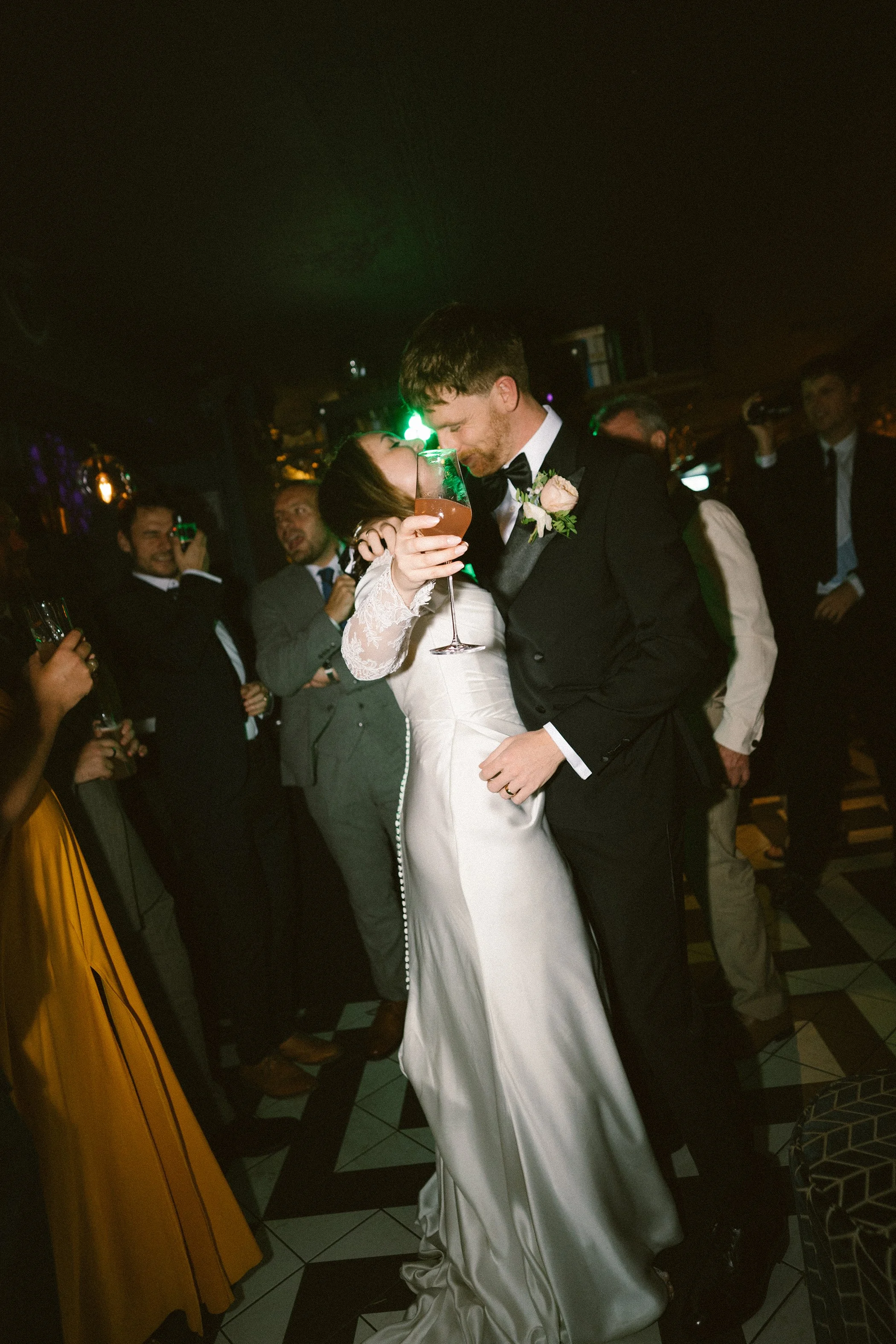 A newlywed couple, the bride in a white gown and the groom in a black tuxedo, sharing a kiss and holding a drink at their wedding reception, surrounded by guests in formal attire.