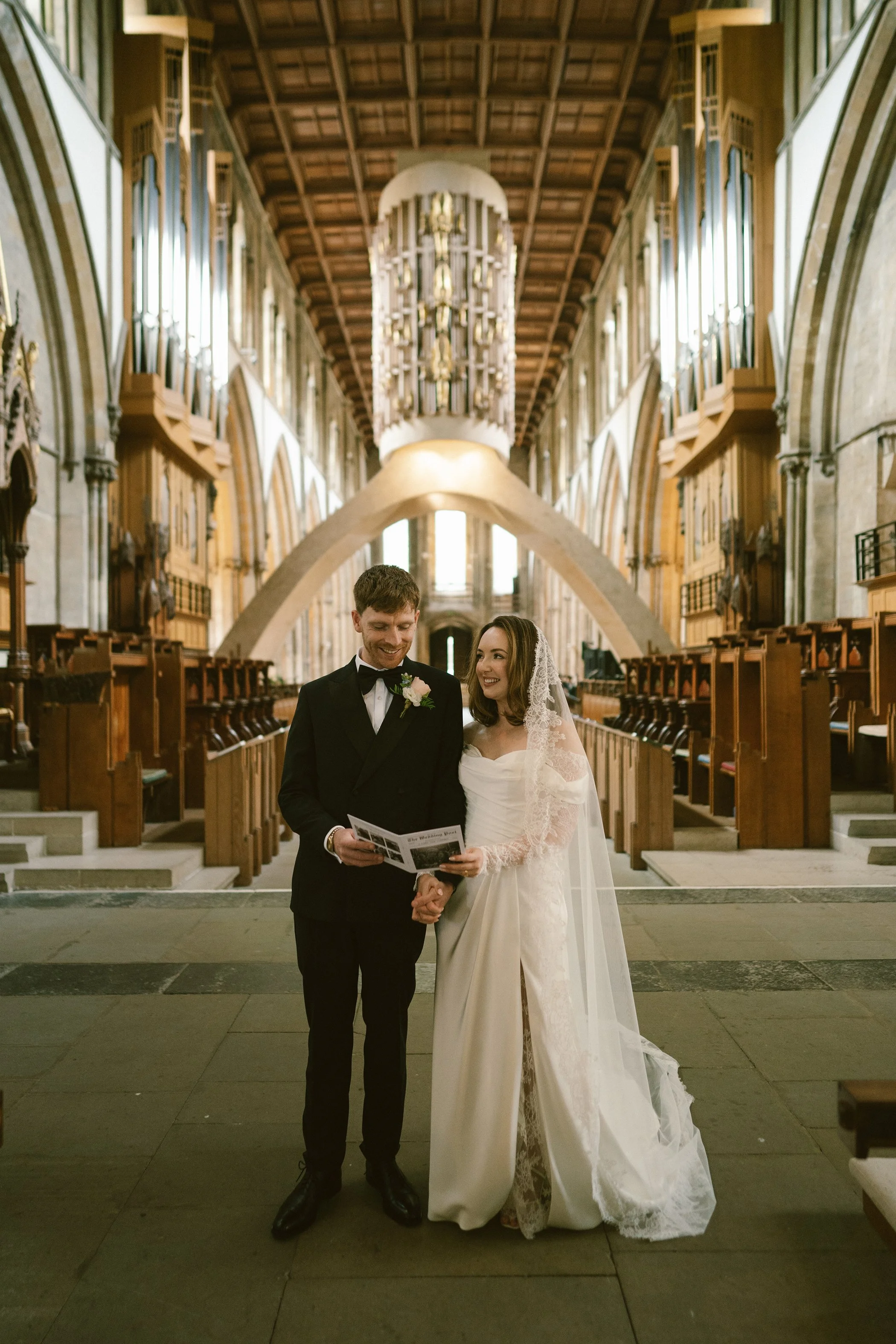A bride and groom standing together in a church during their wedding ceremony, holding hands and smiling while looking at each other.