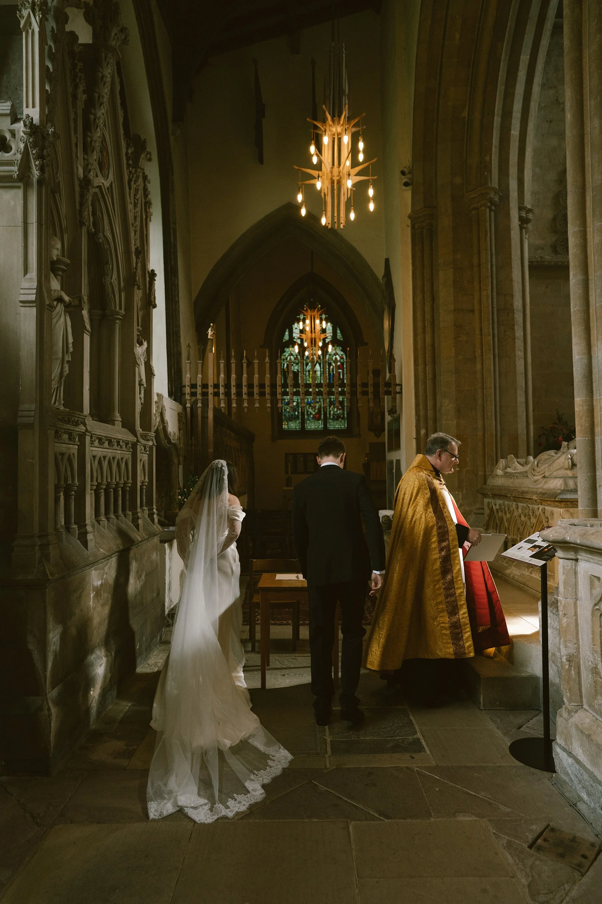 A church interior during a wedding ceremony, with a bride in a long white wedding gown and veil, and a priest in gold vestments, standing near religious statues and stained glass windows, illuminated by a chandelier.