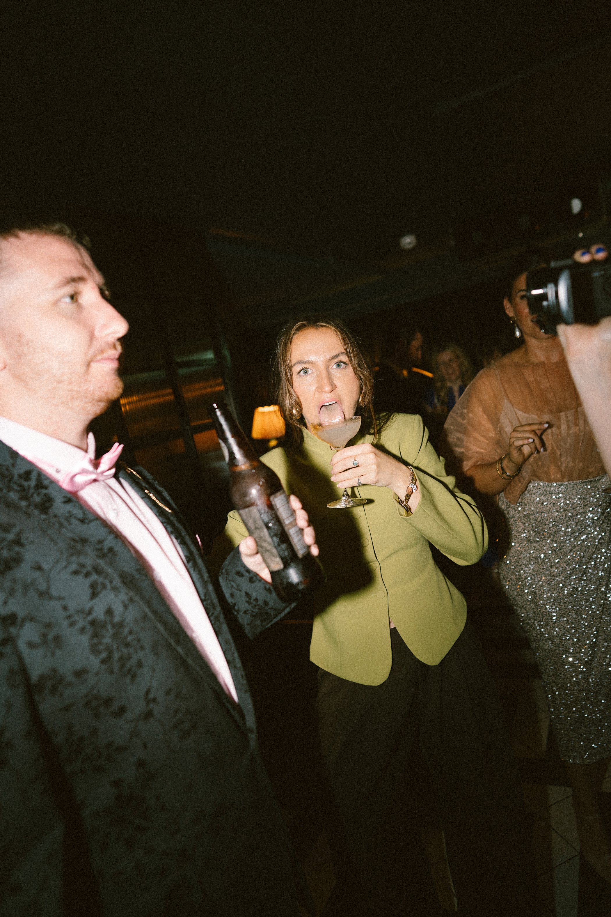 People socializing at an indoor event, including a man in a tuxedo with a pink bow tie, a woman in a yellow blazer holding a drink, and a woman in a glittery dress taking a photo with a camera.
