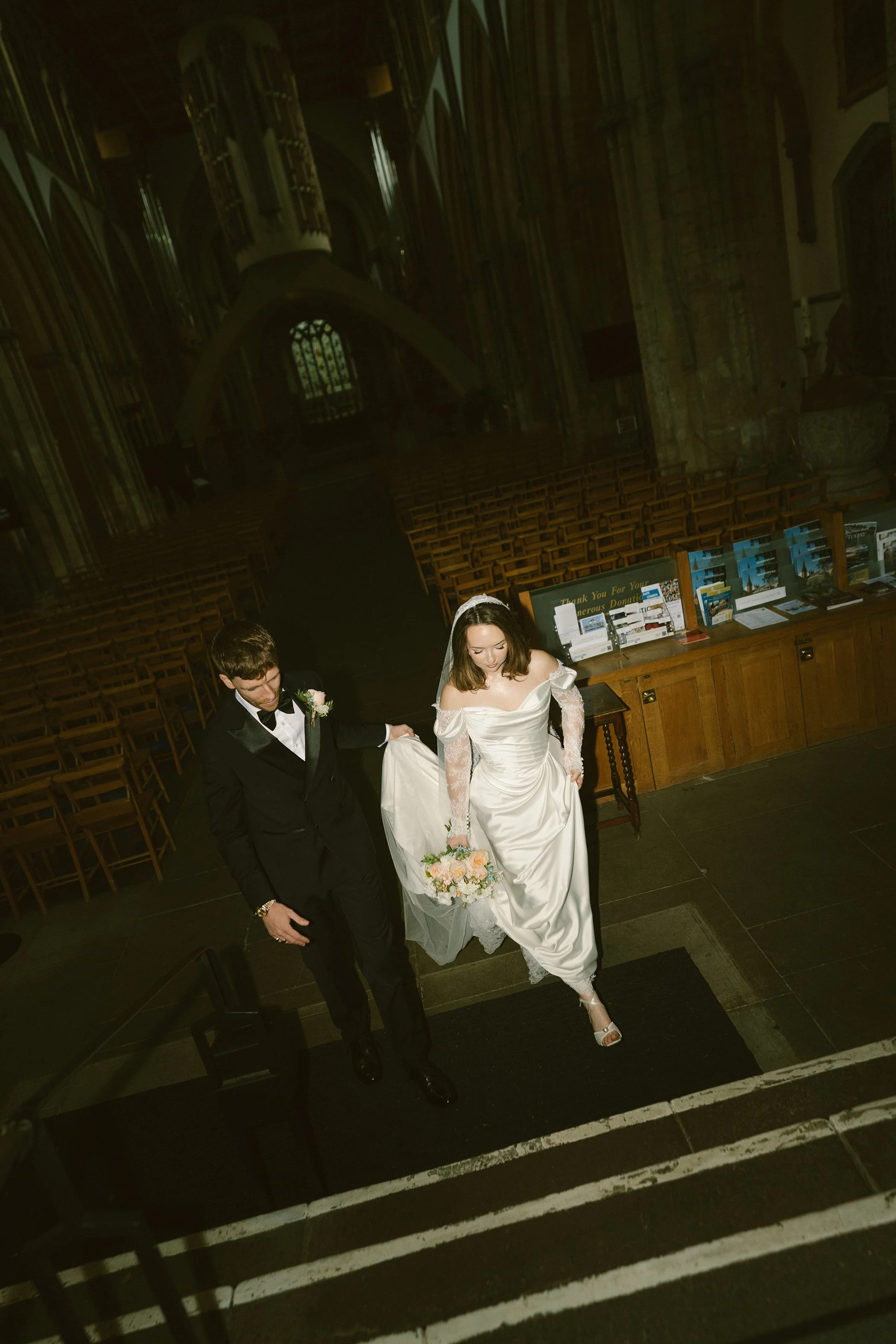 A bride and groom walking up a staircase inside a church. The bride wears a white wedding gown and the groom is in a black tuxedo. The bride holds a bouquet of flowers and there are empty chairs and informational pamphlets in the background.