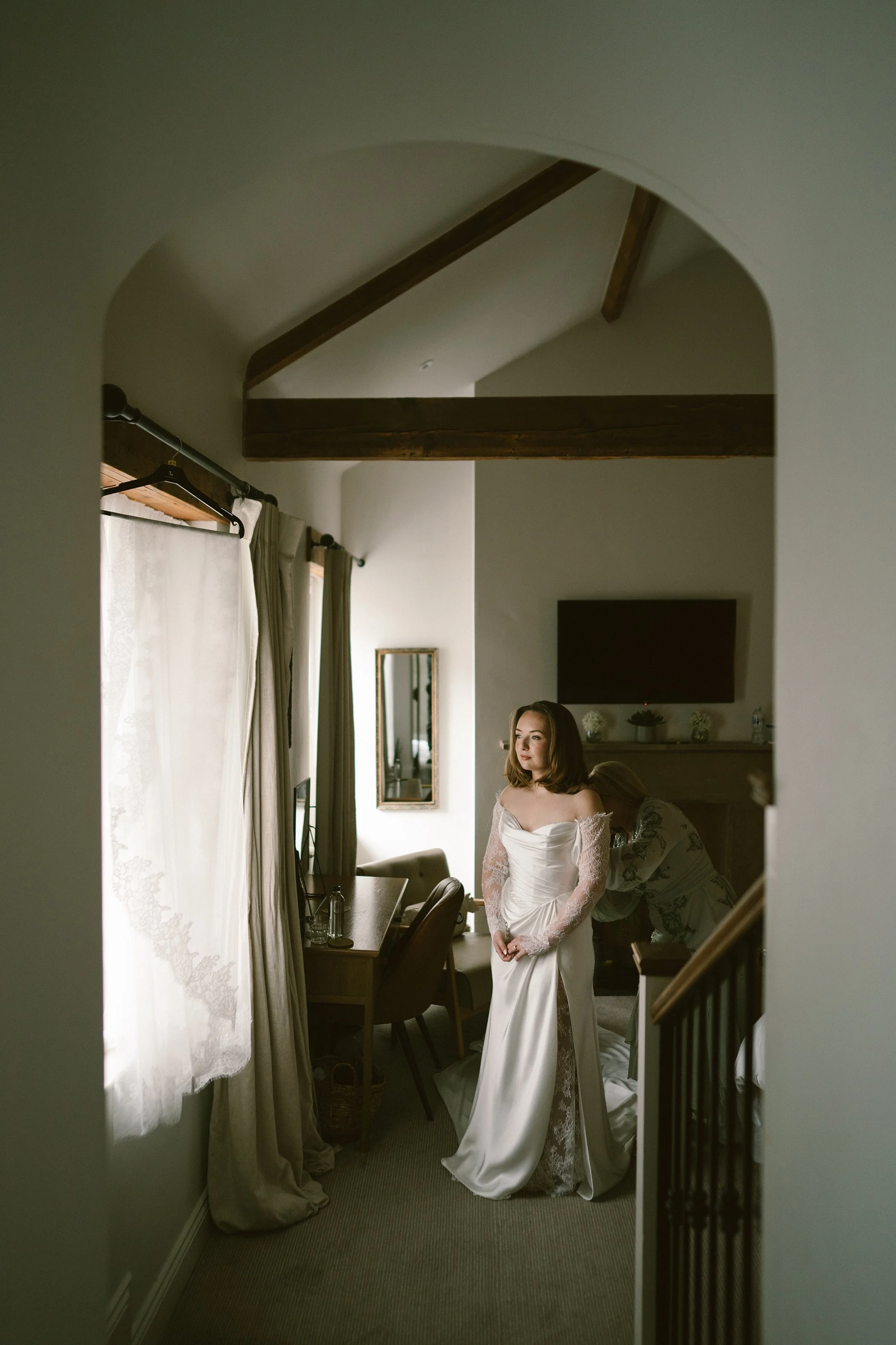 A woman in a white satin wedding dress with lace sleeves standing in a room, being assisted by another person behind her, with a mirror, curtains, and a wall-mounted TV in the background.