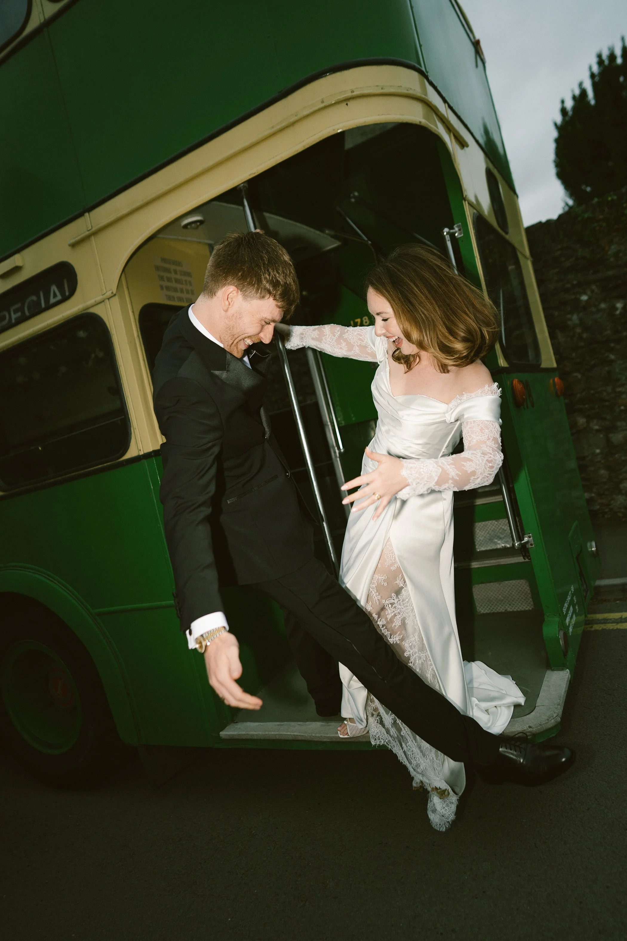 A bride and groom are joyfully stepping off a vintage green and cream bus, smiling and holding hands, with the bride in a white lace wedding dress and the groom in a black suit.