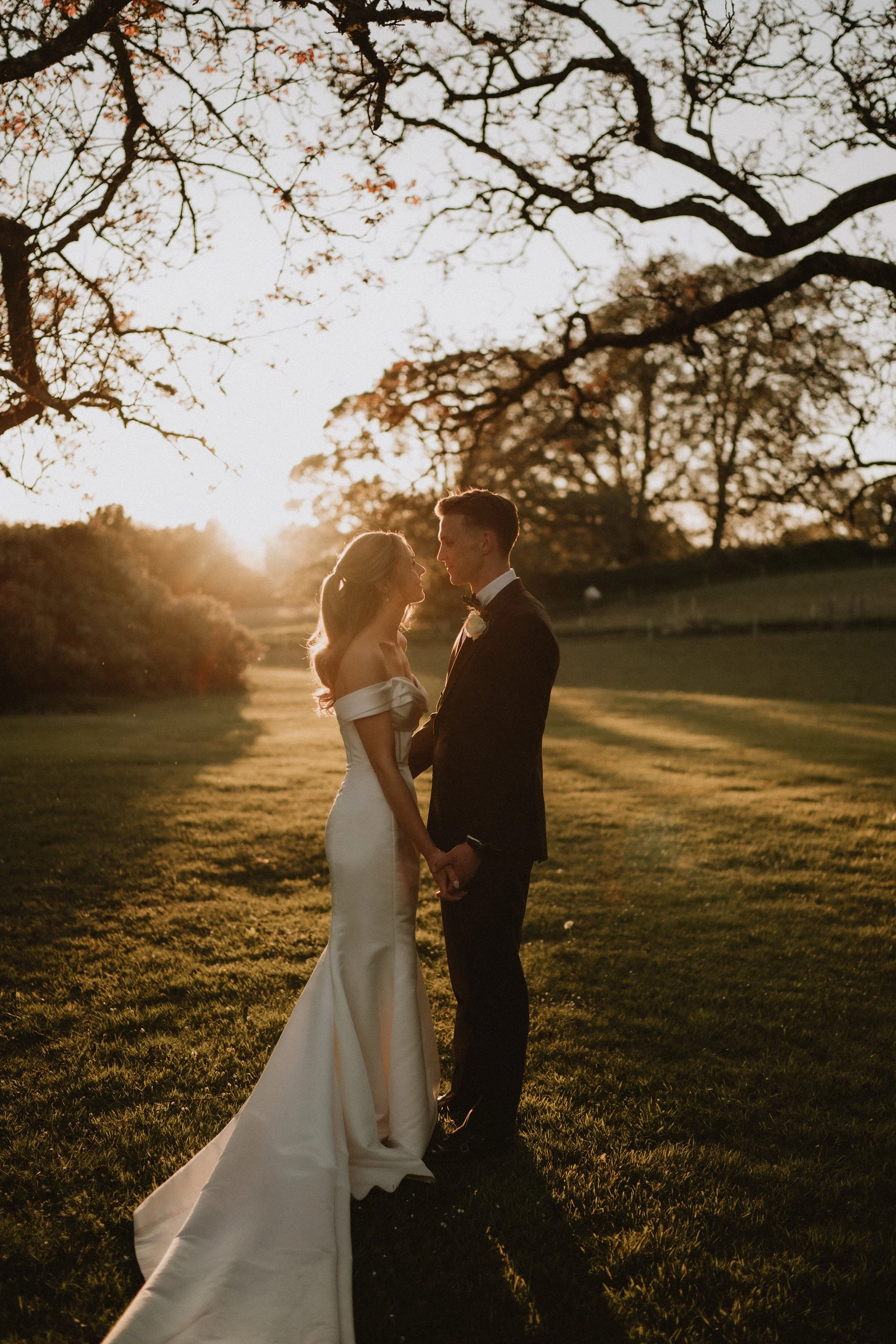 A bride and groom holding hands and looking at each other at sunset in a grassy field with trees.
