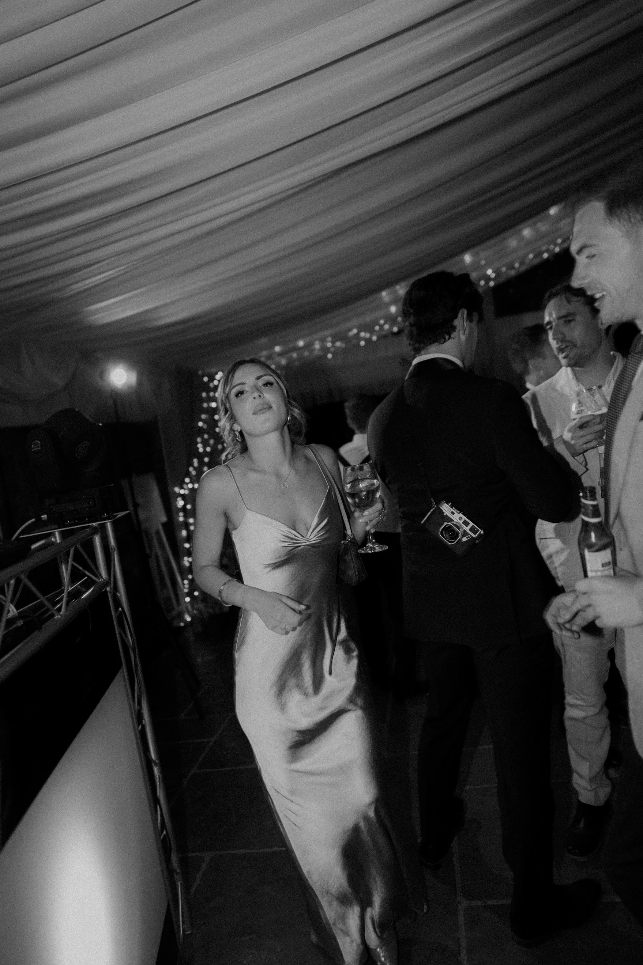 Black and white photo of a woman in a long satin dress holding a glass of wine at a party or wedding reception, surrounded by other people socializing.