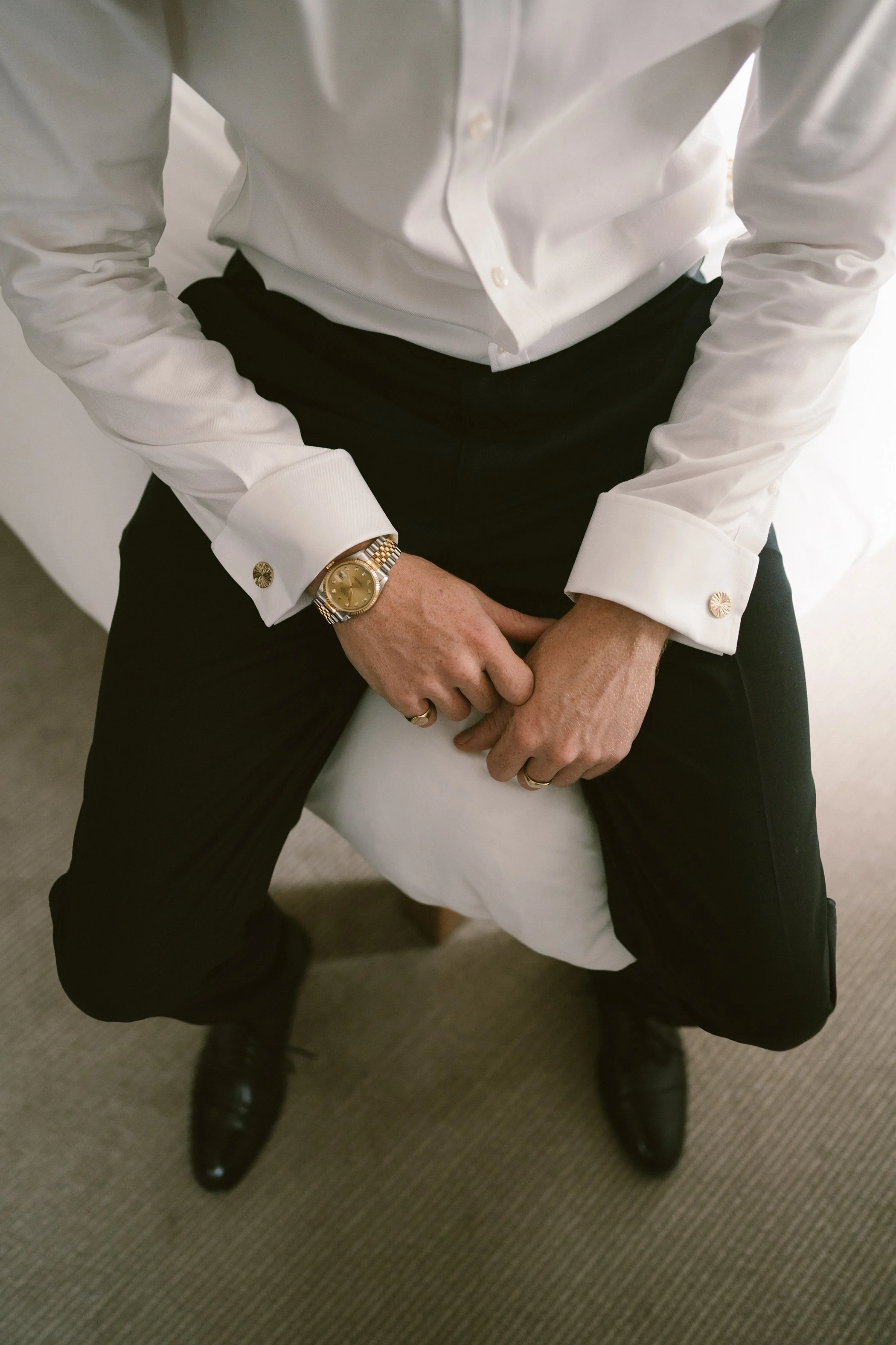 Person sitting on a white chair, wearing a white dress shirt with gold cufflinks, black pants, black shoes, and a gold wristwatch. Hands are clasped in lap.