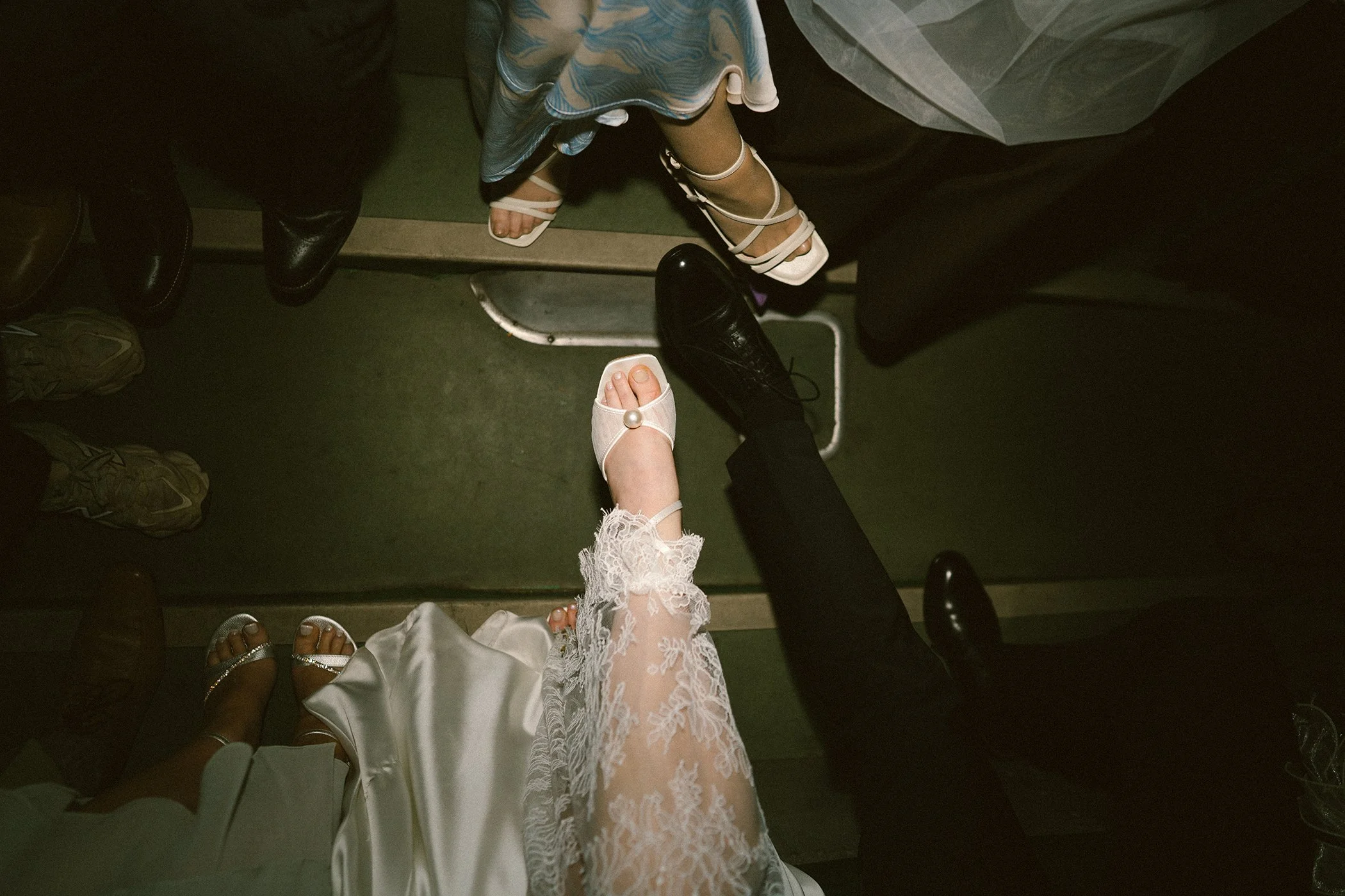 View of people sitting in line, with focus on their feet. Some are wearing sandals or sneakers, others are wearing formal shoes. The image is taken from above, showing various styles of footwear on a green floor.