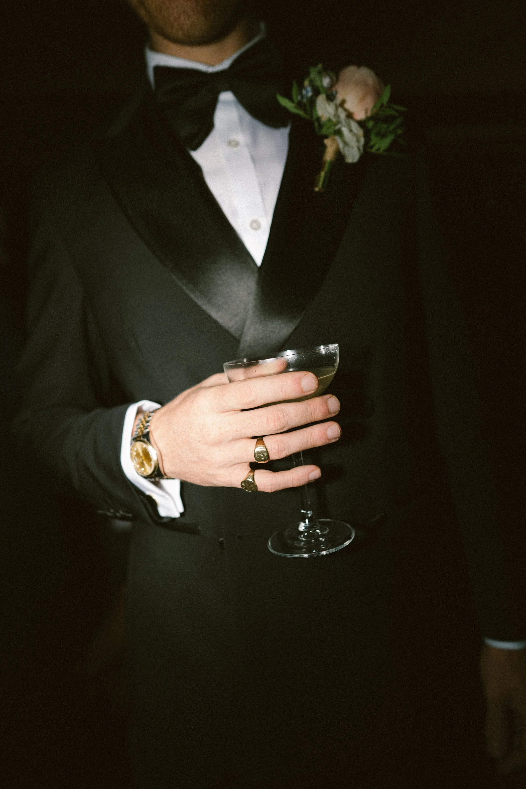 A man in a formal tuxedo with a black bow tie and white shirt holds a champagne glass in his hand. He is wearing a gold wristwatch and rings on his fingers, and has a boutonniere with white flowers and greenery on his lapel.