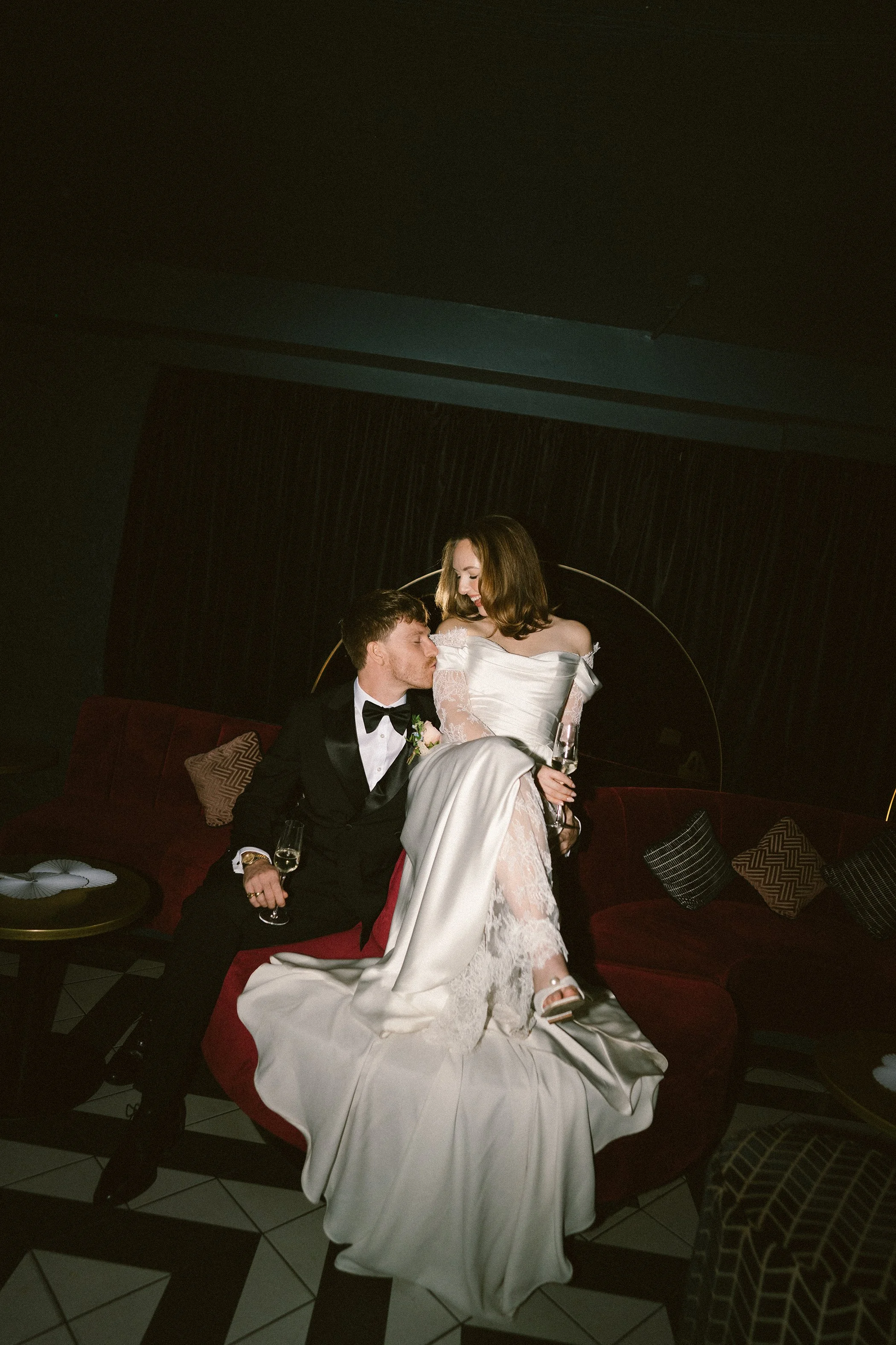 A couple at their wedding reception, with the bride in a white gown sitting on the groom's lap on a red velvet sofa, both holding champagne glasses, in a room with dark curtains and patterned pillows.
