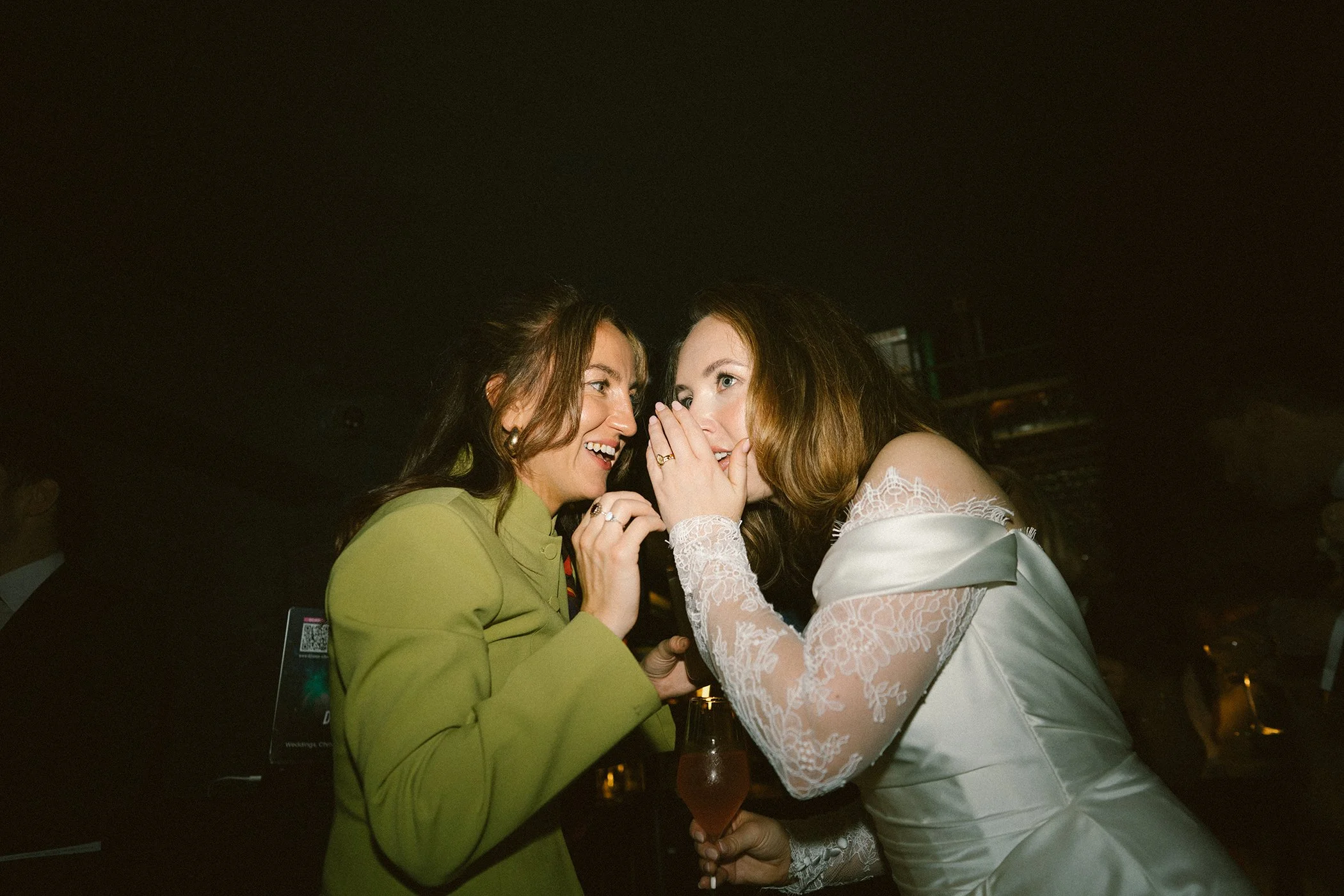 Two women whispering to each other at night in a dark setting, one wearing a green jacket and the other in a white lace dress.