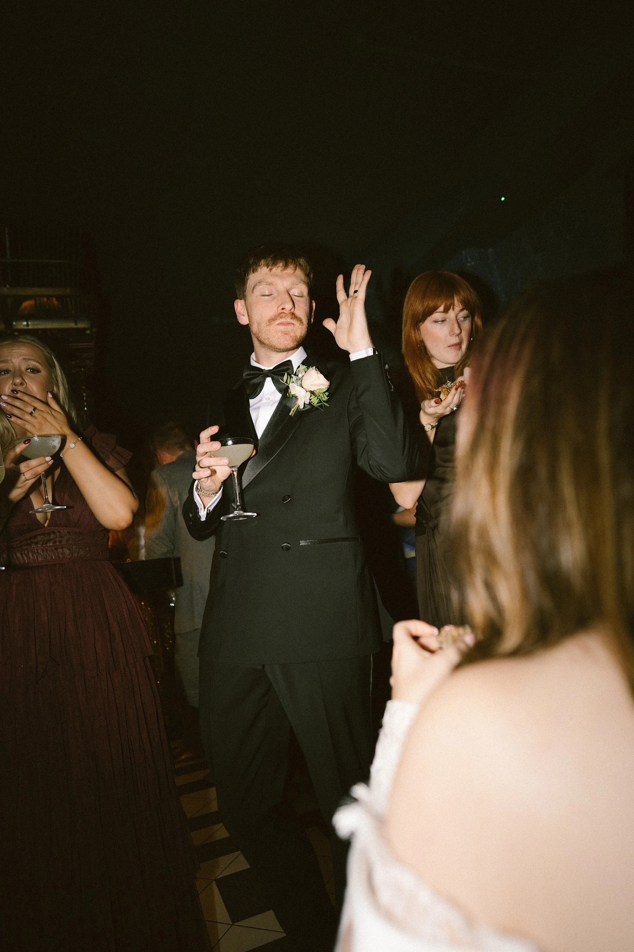 A man in a black tuxedo, holding a glass, with his eyes closed and one hand raised, appears to be at a formal event or celebration with women and other guests around him.