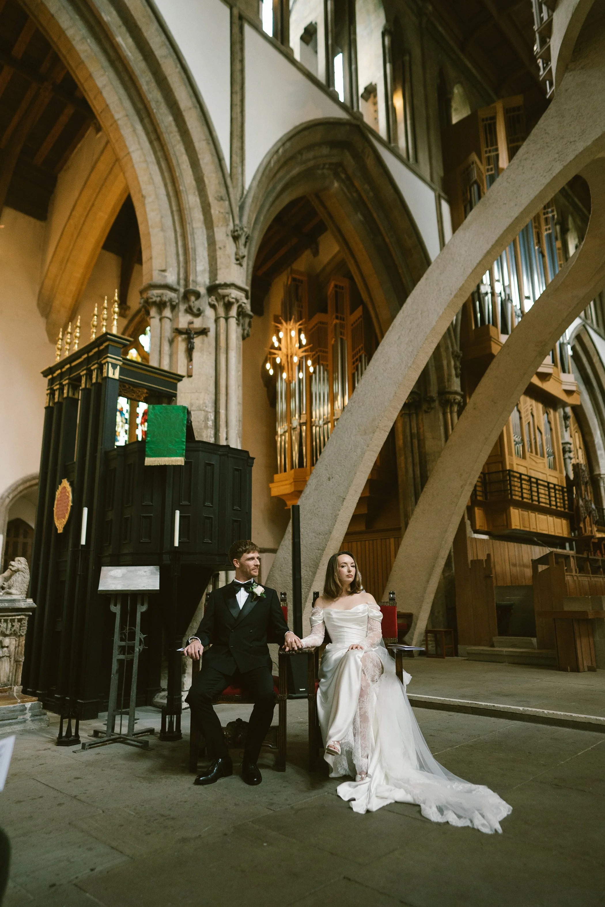 A bride and groom sitting together on a throne-like chair in a church with Gothic architecture. The groom is wearing a tuxedo and the bride is in a white wedding dress. They are holding hands and looking in opposite directions.