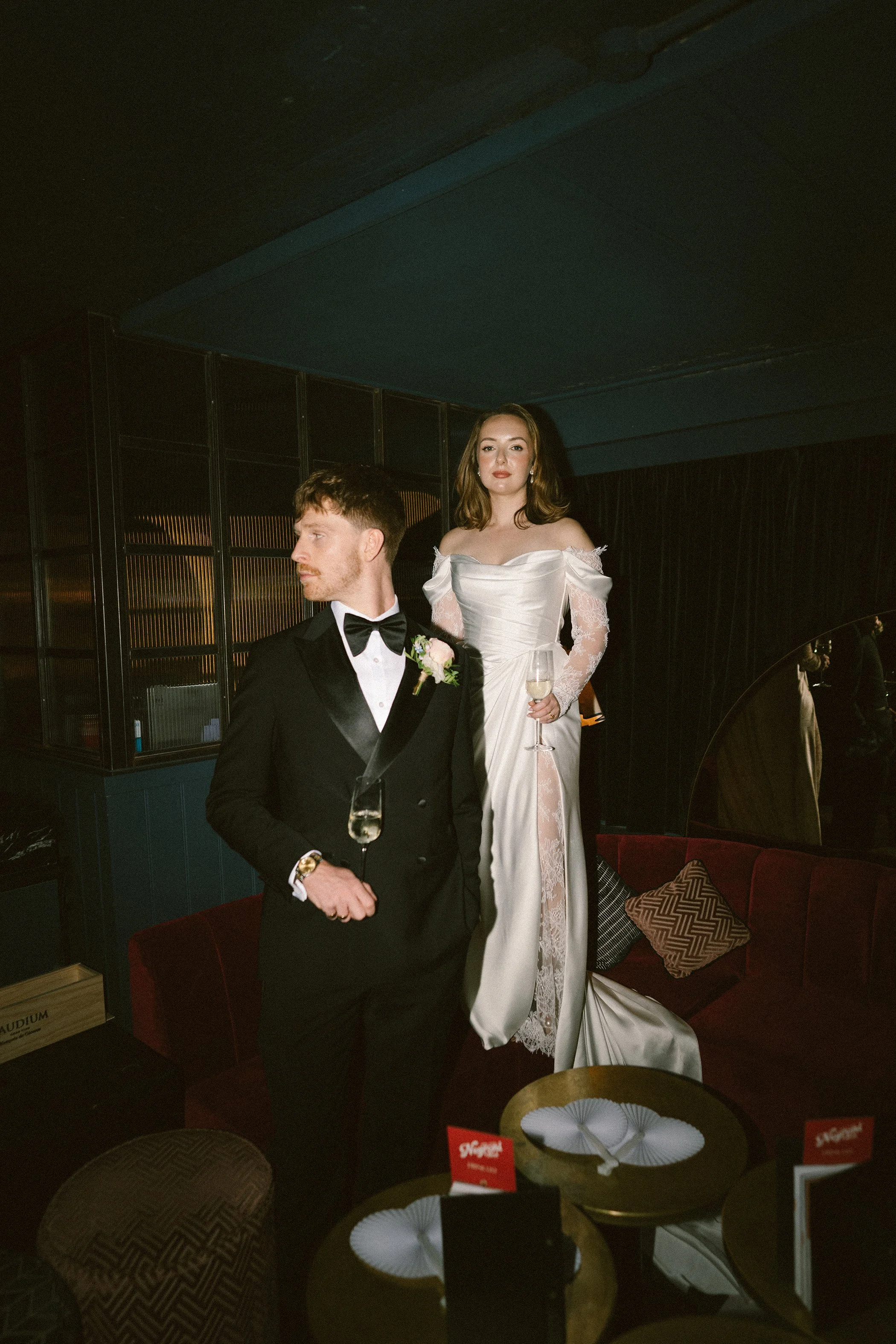 A man in a tuxedo and a woman in an elegant wedding dress at an indoor event. The woman is holding a glass of champagne and standing behind the man. The scene appears to be a celebration or wedding reception.